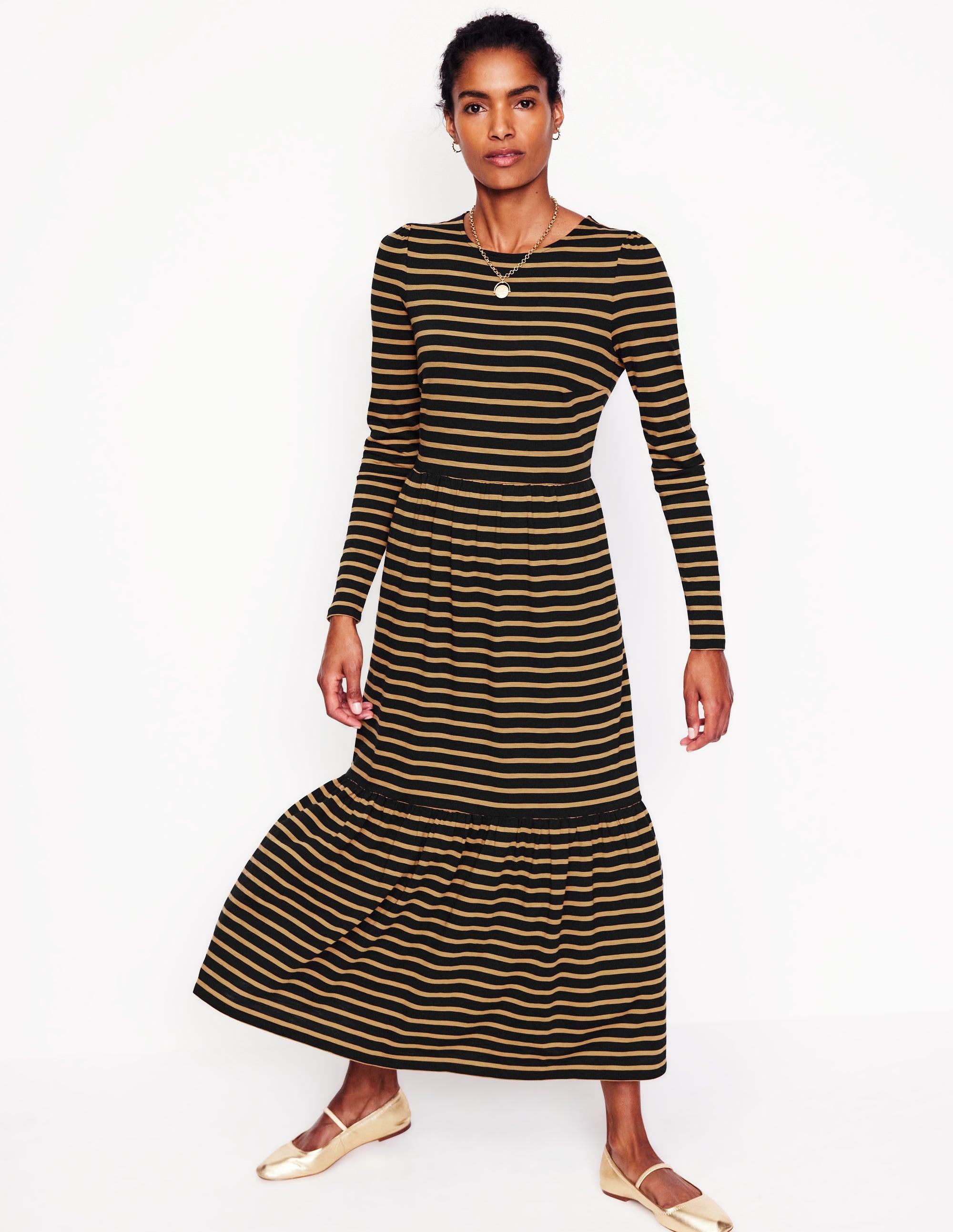 A woman wearing a long sleeve jersey dress with camel and black horizontal stripes, fitted bodice, and gathered tiers, paired with beige ballet flats.