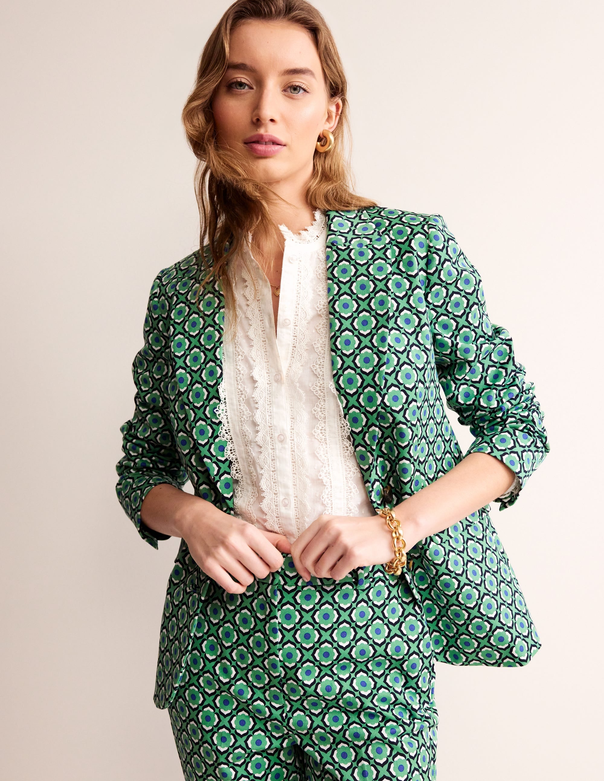 A woman wearing a Ming Green patterned blazer with a geometric design, revere collar, and contrast piping, paired with matching patterned trousers and a white lace blouse.