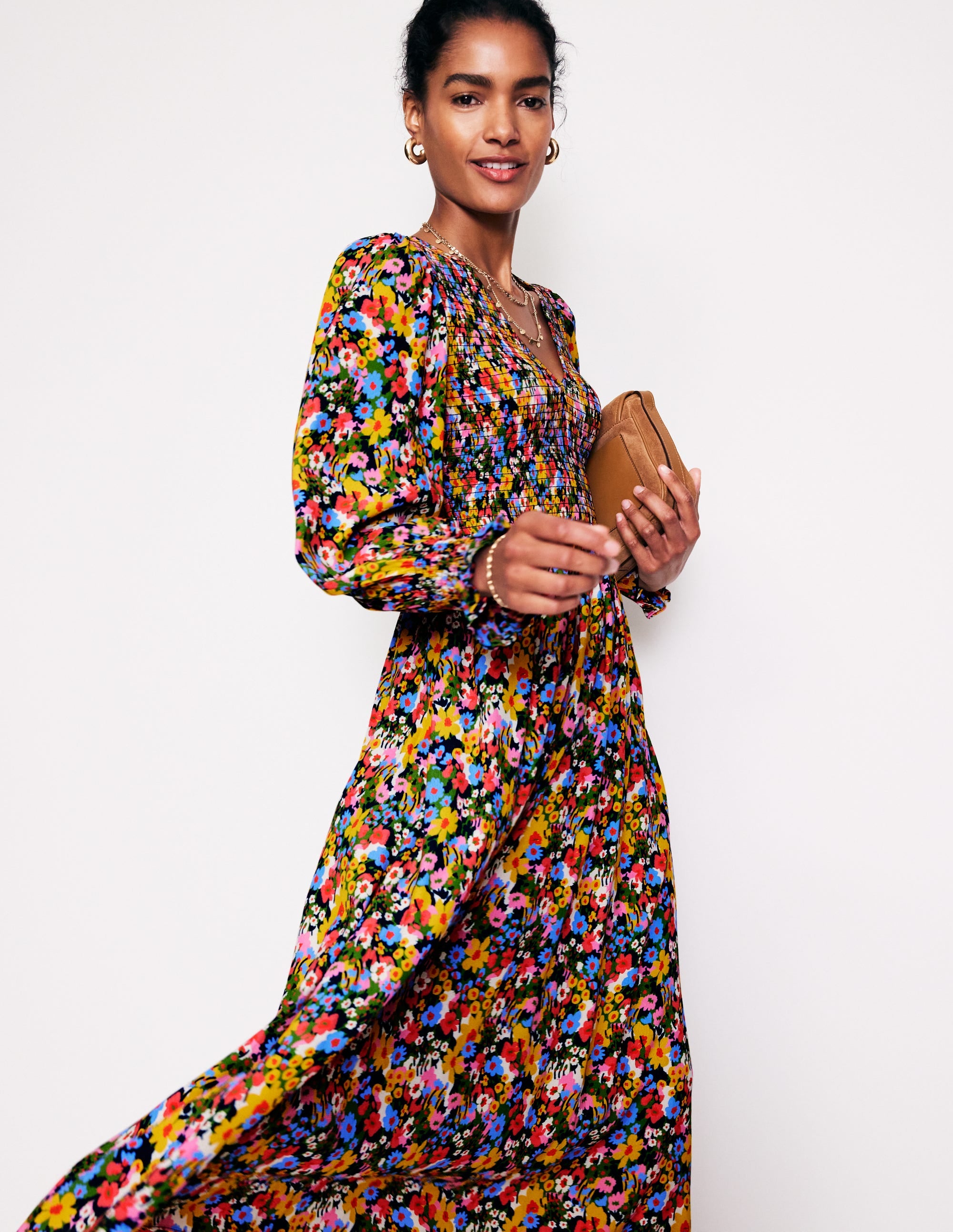 A woman wearing a colorful floral midi dress with long sleeves, smocking details, and side pockets, holding a tan clutch.