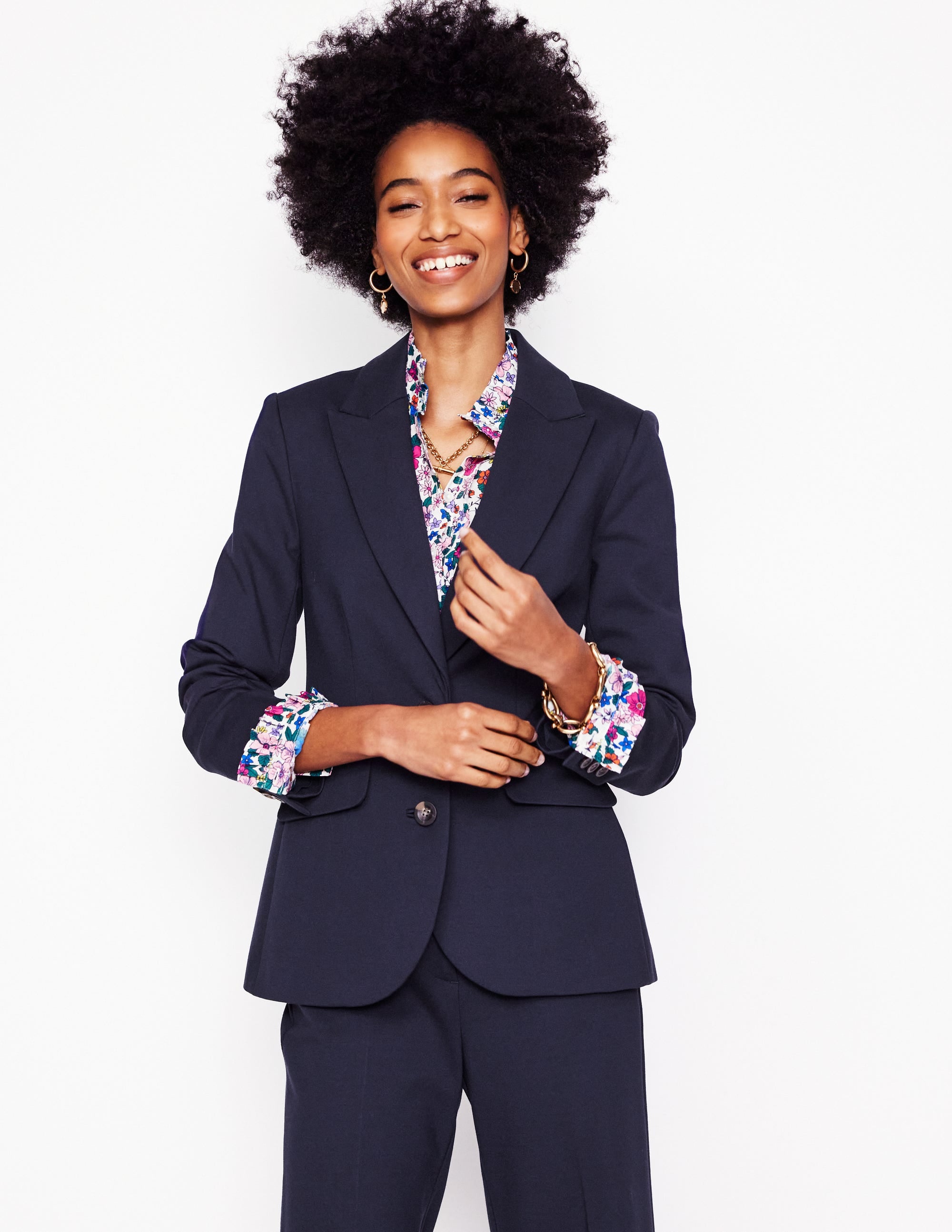 A woman wearing a navy blazer with a floral shirt underneath, smiling and standing against a plain white background.