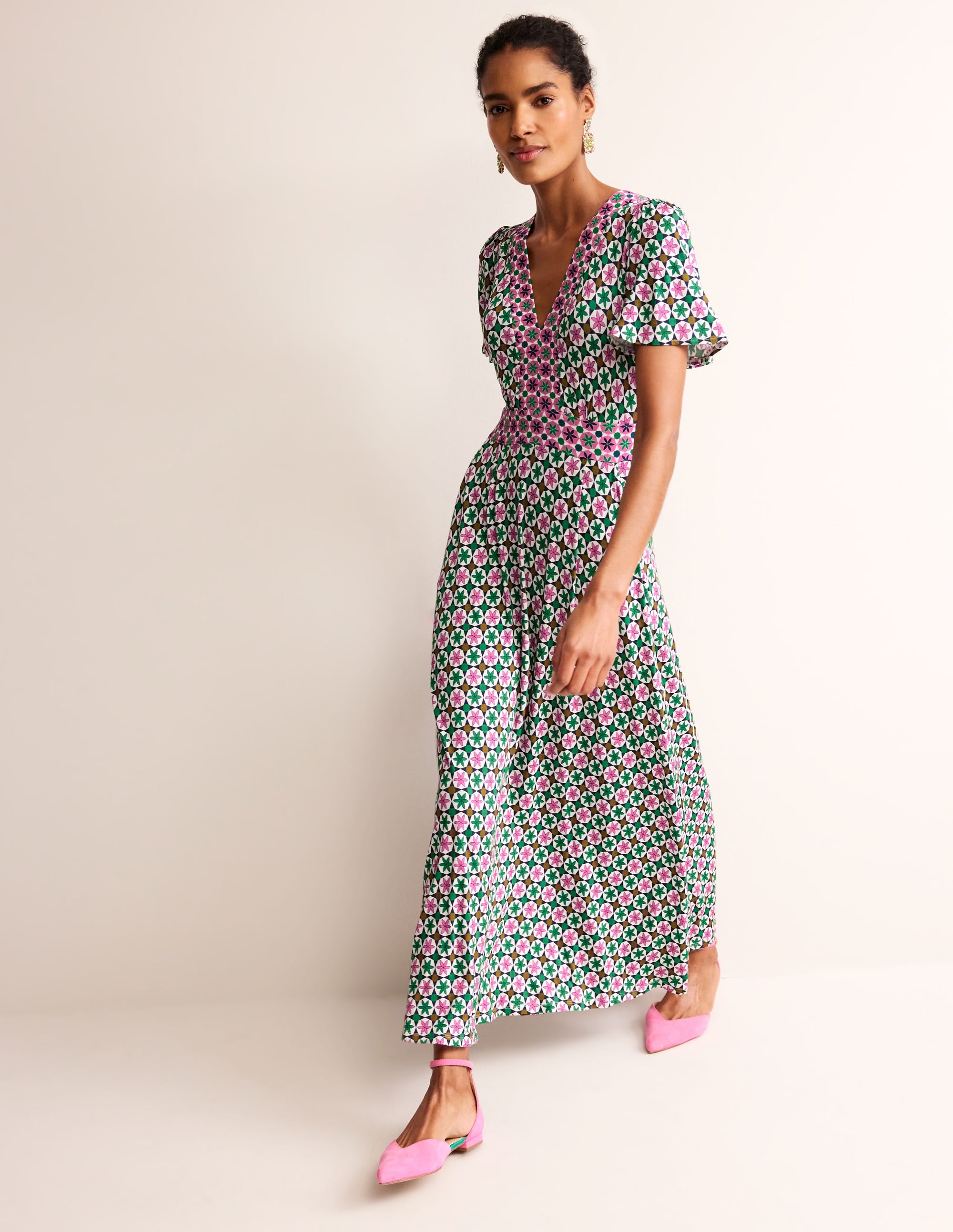 A woman wearing a maxi dress with a multicolour botanical tile print, V-neckline, flutter sleeves, and side pockets.