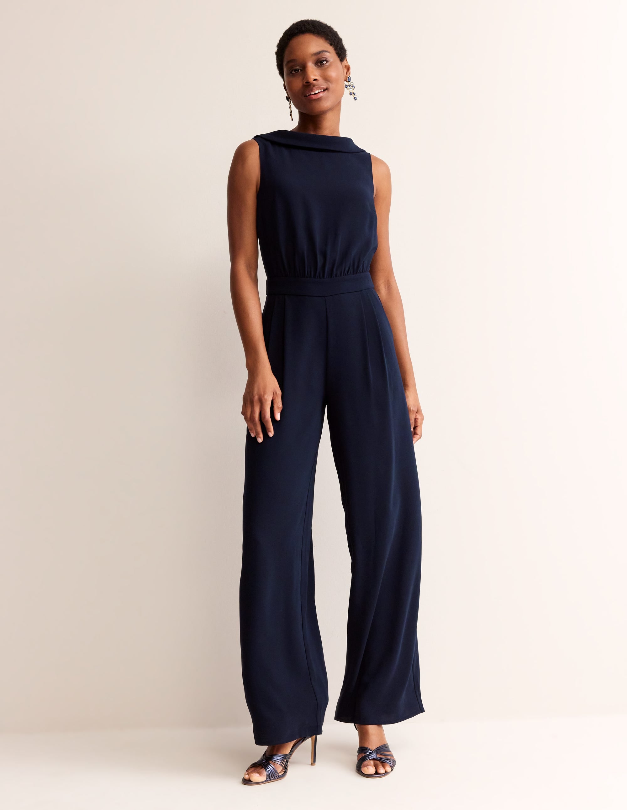 A woman wearing a navy jumpsuit with a fitted bodice and wide-leg trousers, sleeveless with a high neckline, standing against a plain background.