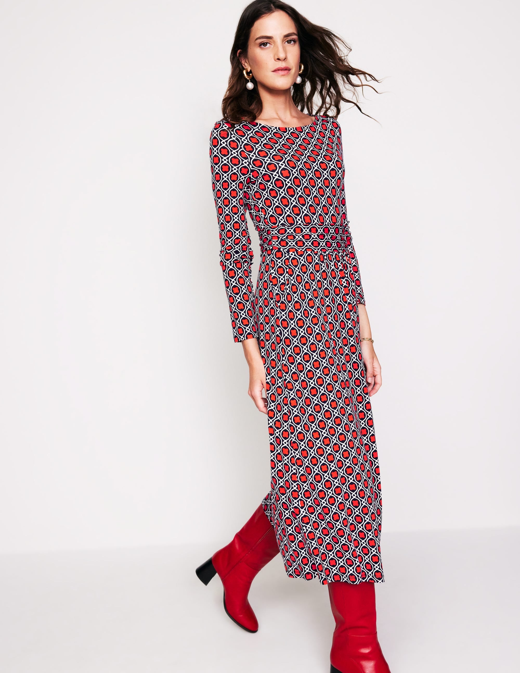 A woman wearing a long-sleeve midi dress with a geometric pattern in red, black, and white, paired with red knee-high boots.
