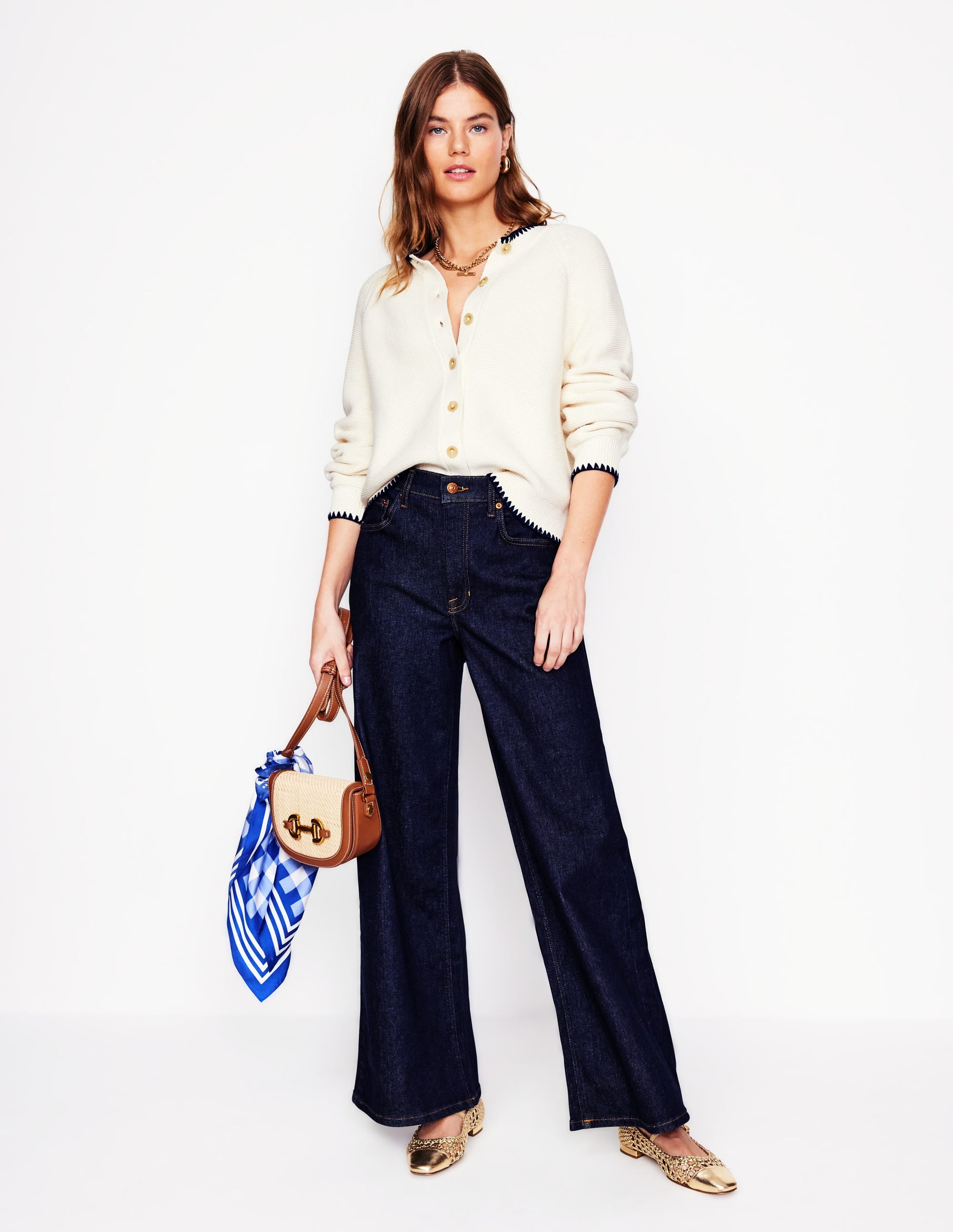A woman wearing high-rise wide-leg jeans in indigo rinse, paired with a cream sweater, holding a handbag, and wearing gold shoes.