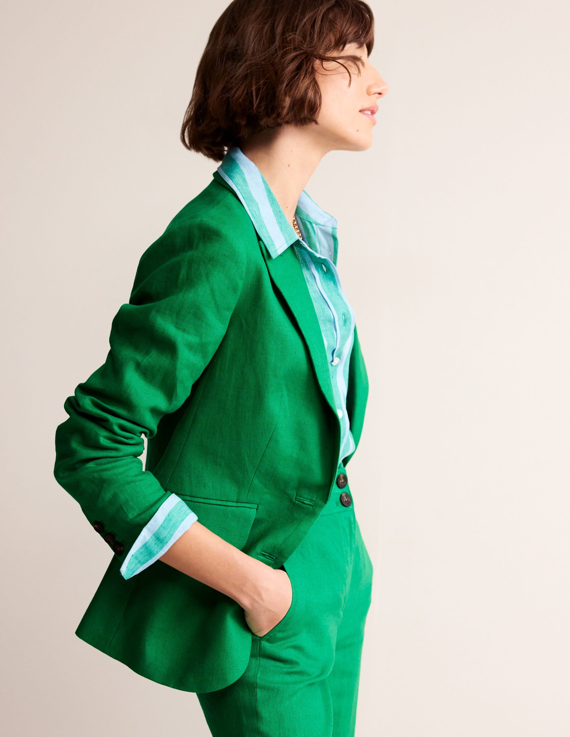 A woman wearing a green linen blazer with a revere collar, contrast piping, and welt pockets, styled with a matching green shirt underneath.