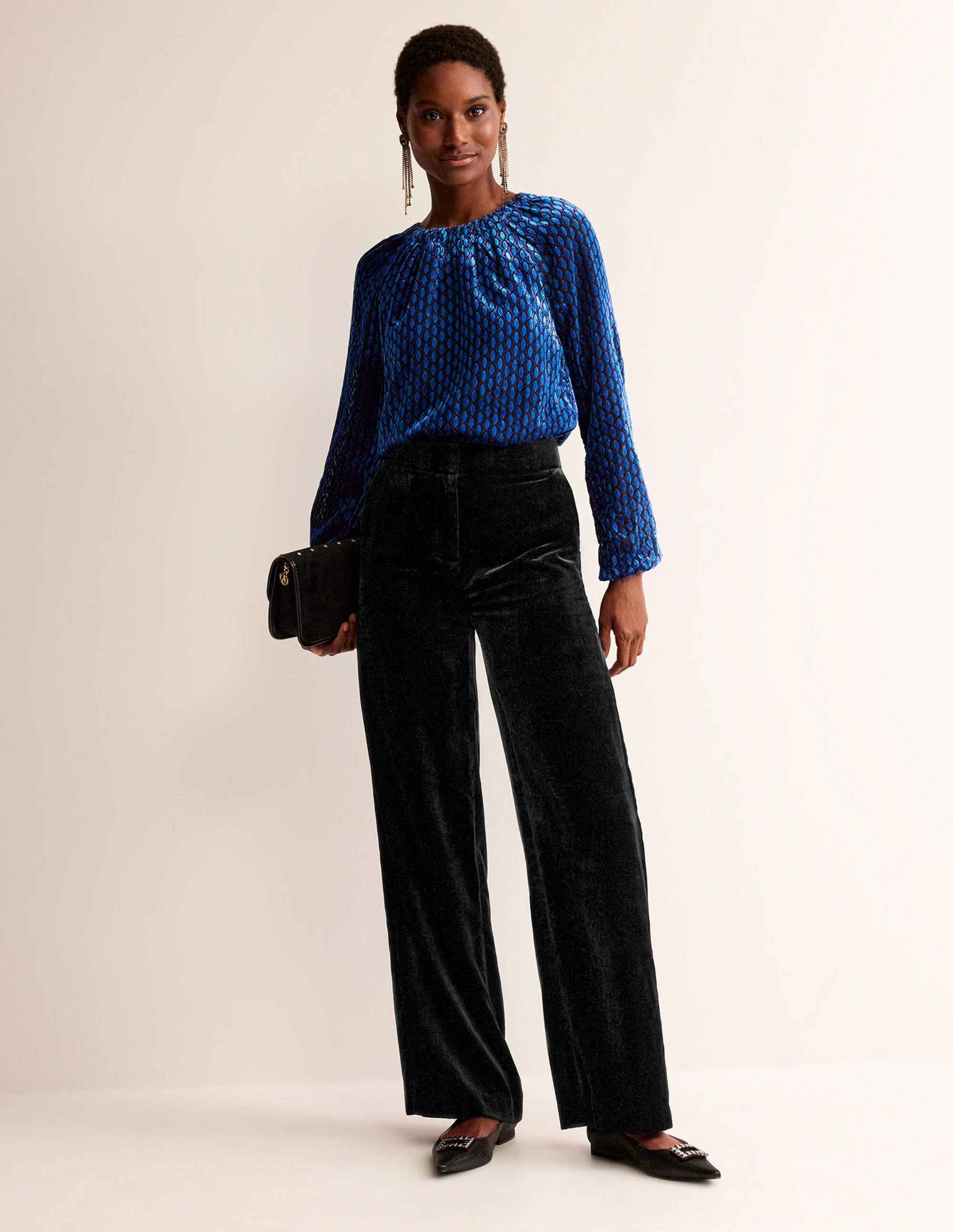 A woman wearing black velvet trousers with a high waist and wide-leg shape, paired with a blue patterned top, holding a black clutch, and wearing black shoes.
