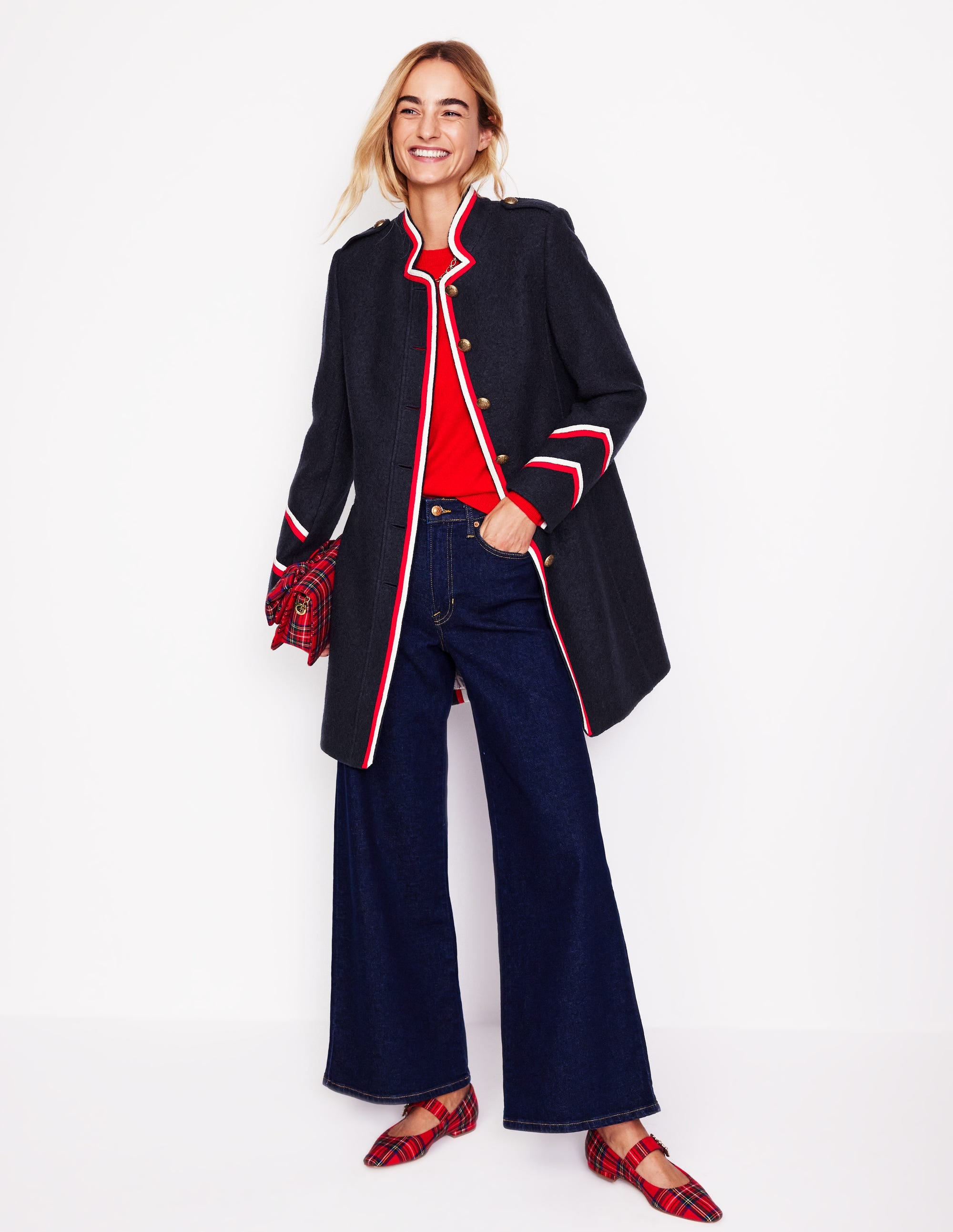 A woman wearing a navy coat with red, white, and navy tipped trim, gold buttons, and a high funnel neck. The coat is knee-length with welt pockets and a straight fit.