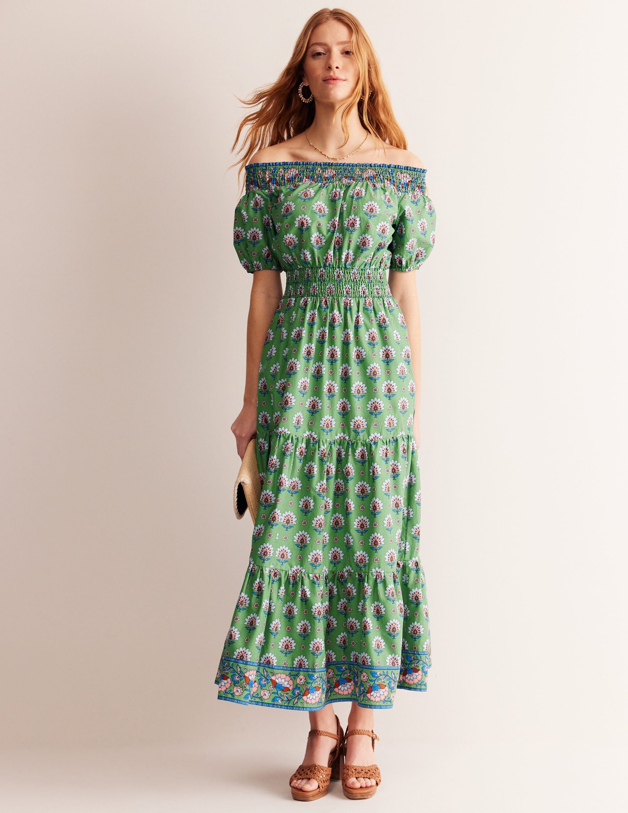 A woman wearing a green floral midi dress with smocked details and a bardot neckline, standing against a plain background.
