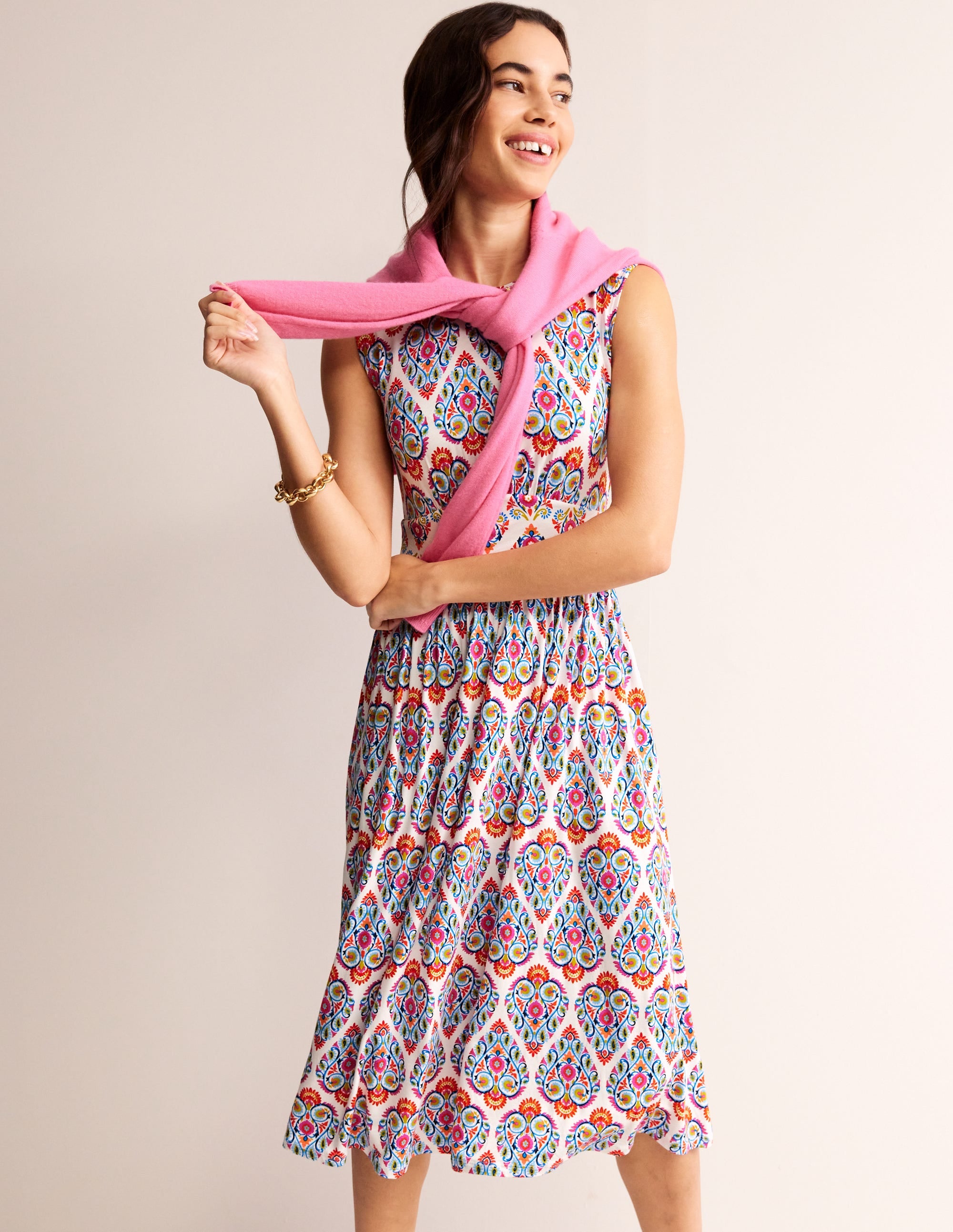 A woman wearing a sleeveless, multicolour floral and geometric print midi dress with a fitted top and contrasting waist panel, holding a pink scarf.