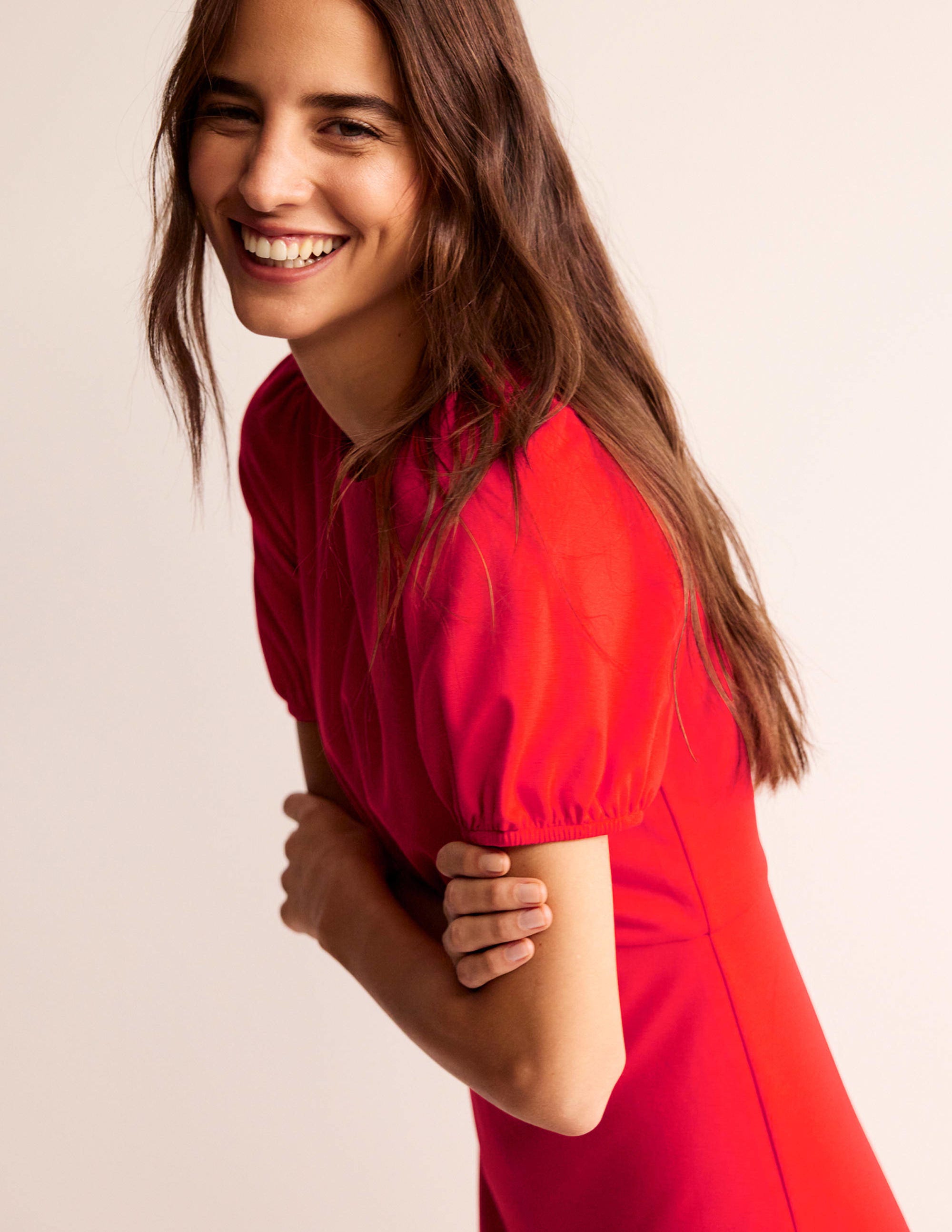 A woman wearing a bright red midi dress with short puffed sleeves and ruching at the waist, smiling.