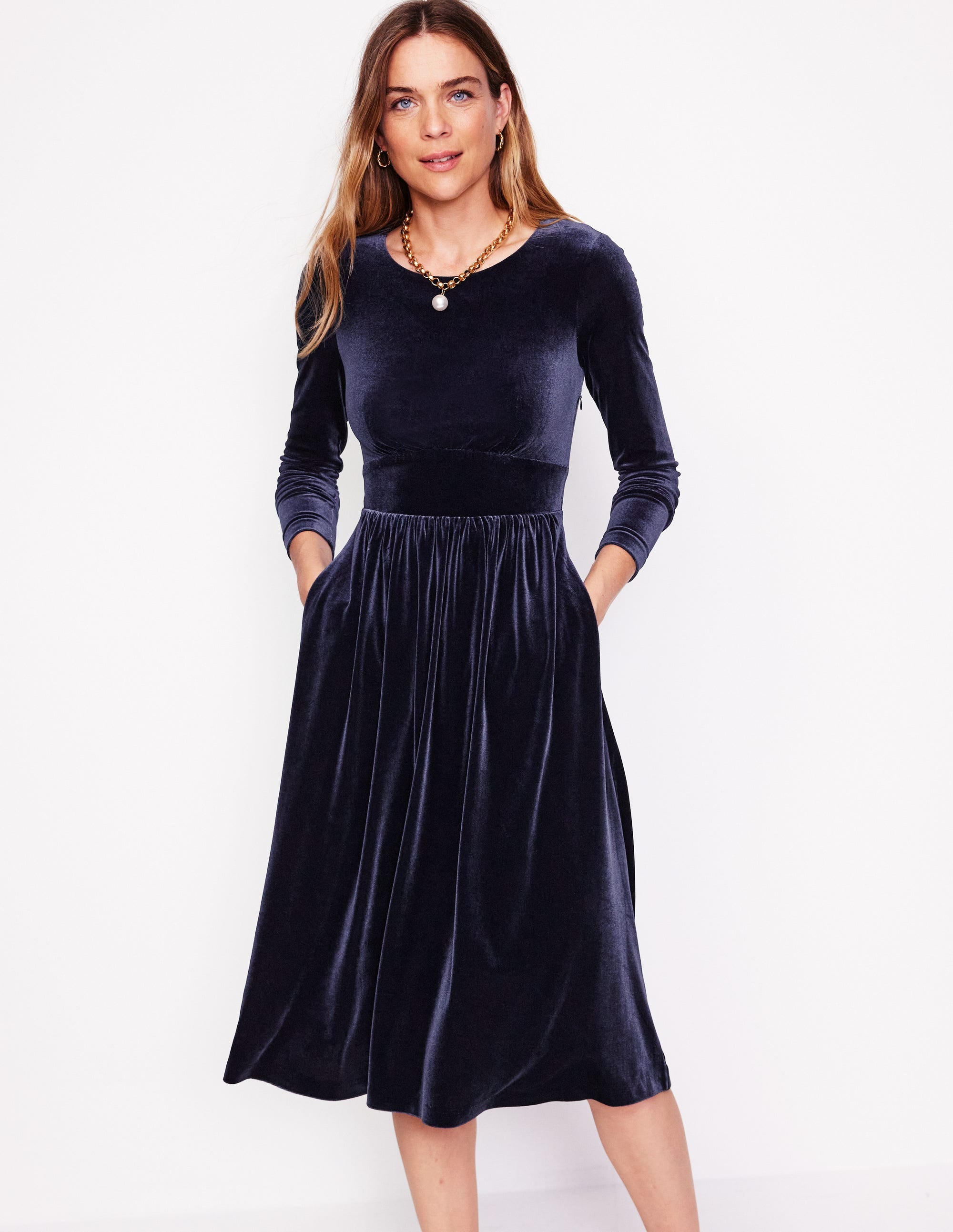 A woman wearing a navy velvet midi dress with long sleeves, a ruched bodice, and a defined waist, standing against a plain white background.