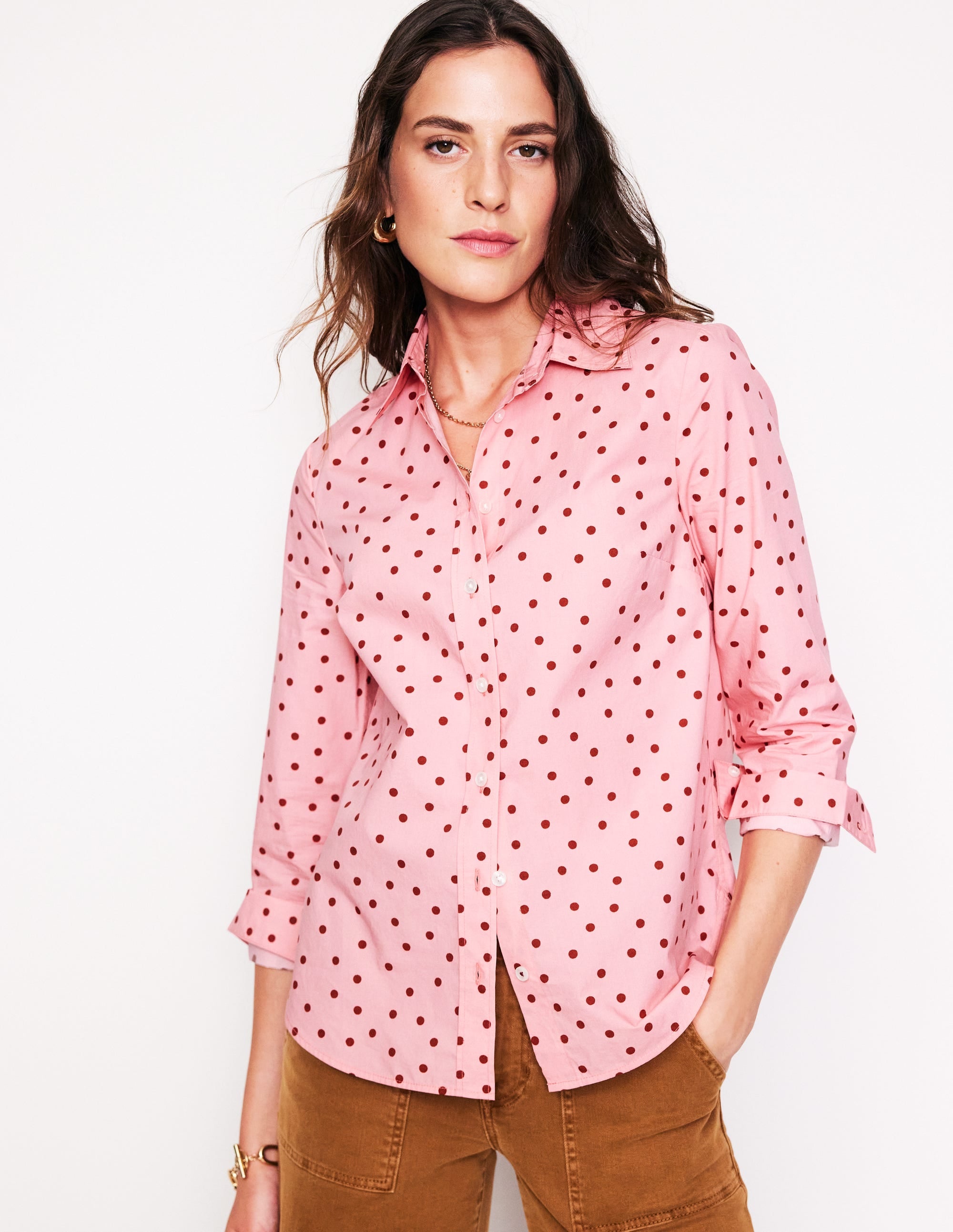 A woman wearing a pink cotton shirt with a sparse spot pattern, long sleeves, and a curved hem, styled casually.