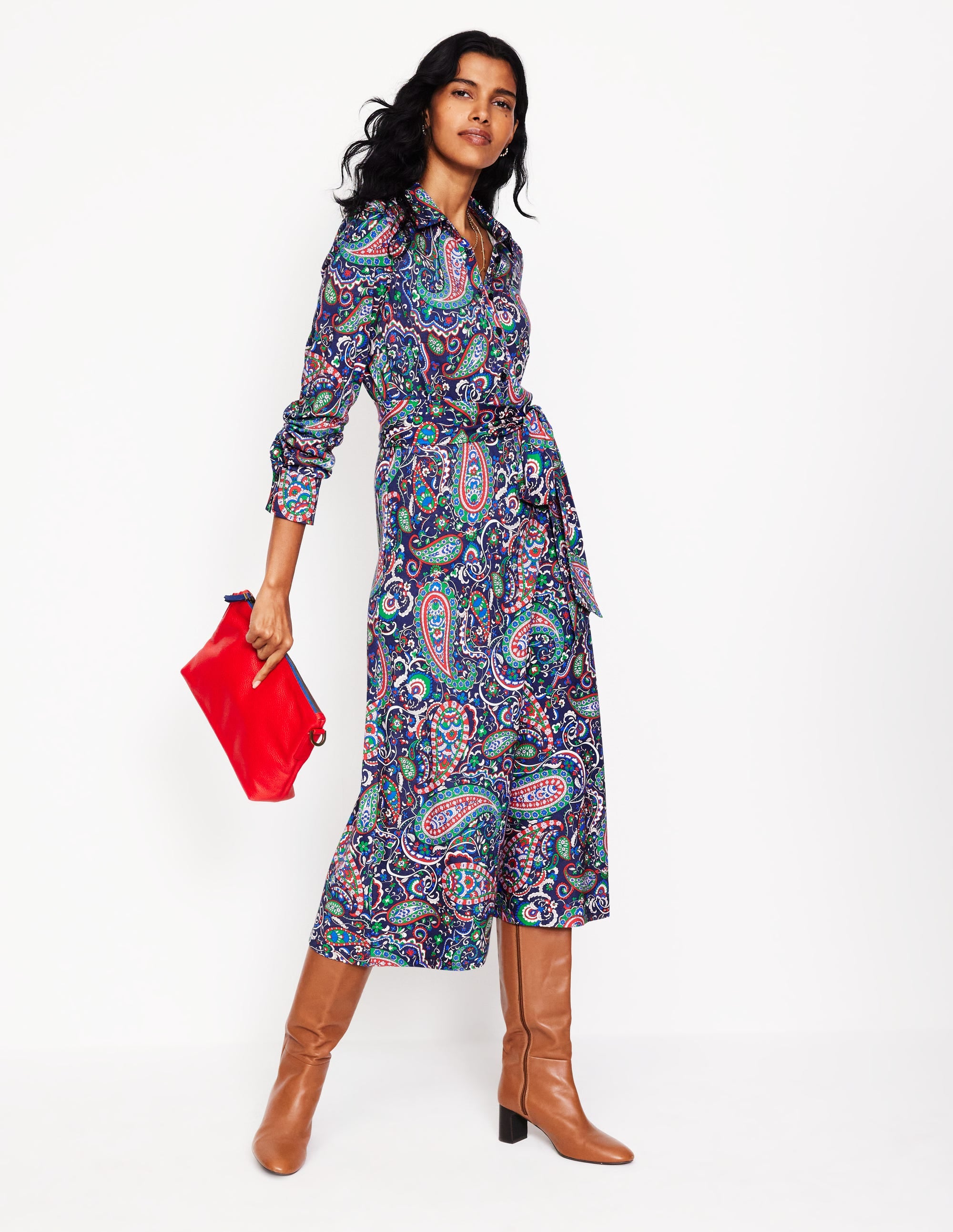 A woman wearing a navy paisley patterned midi shirt dress with a waist belt, paired with brown knee-high boots and holding a red clutch.