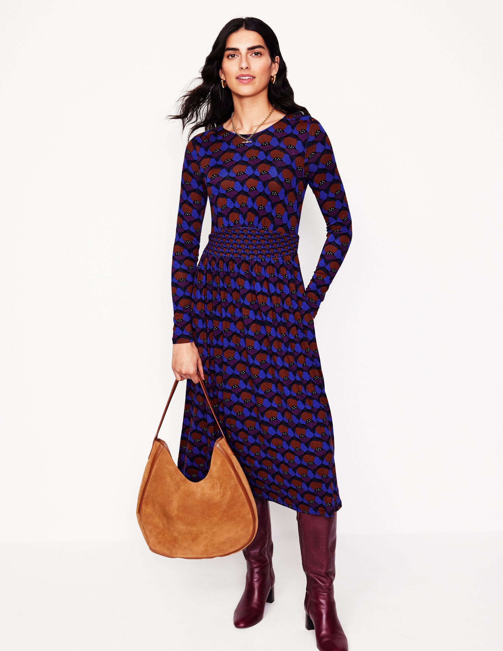 A woman wearing a long sleeve jersey dress with a Damson and Poppy Stamp pattern, fitted waist, and gathered skirt, paired with burgundy boots and a tan bag.