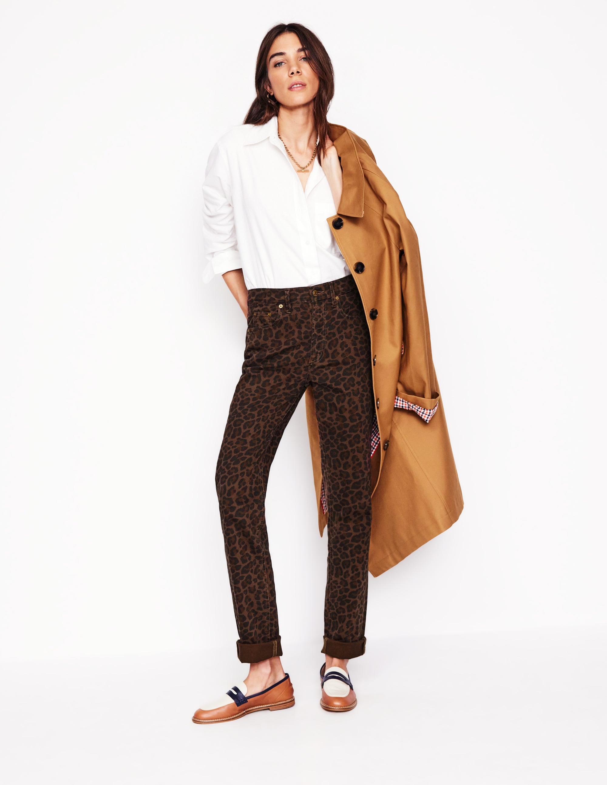 A woman wearing leopard print jeans with rolled-up hems, a white shirt, and a tan coat draped over her shoulder.