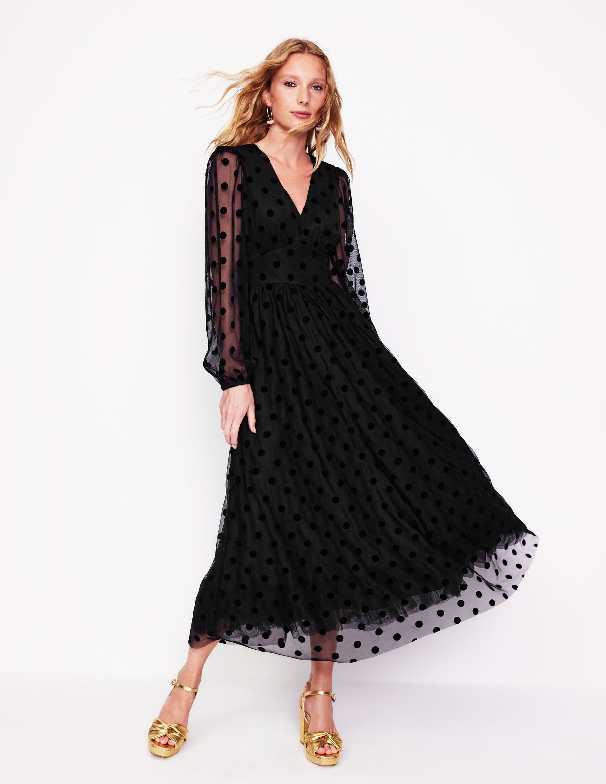 A woman wearing a black midi dress with a layered spotted tulle overlay, puffed long sleeves, and a flared skirt, paired with gold high-heeled sandals.