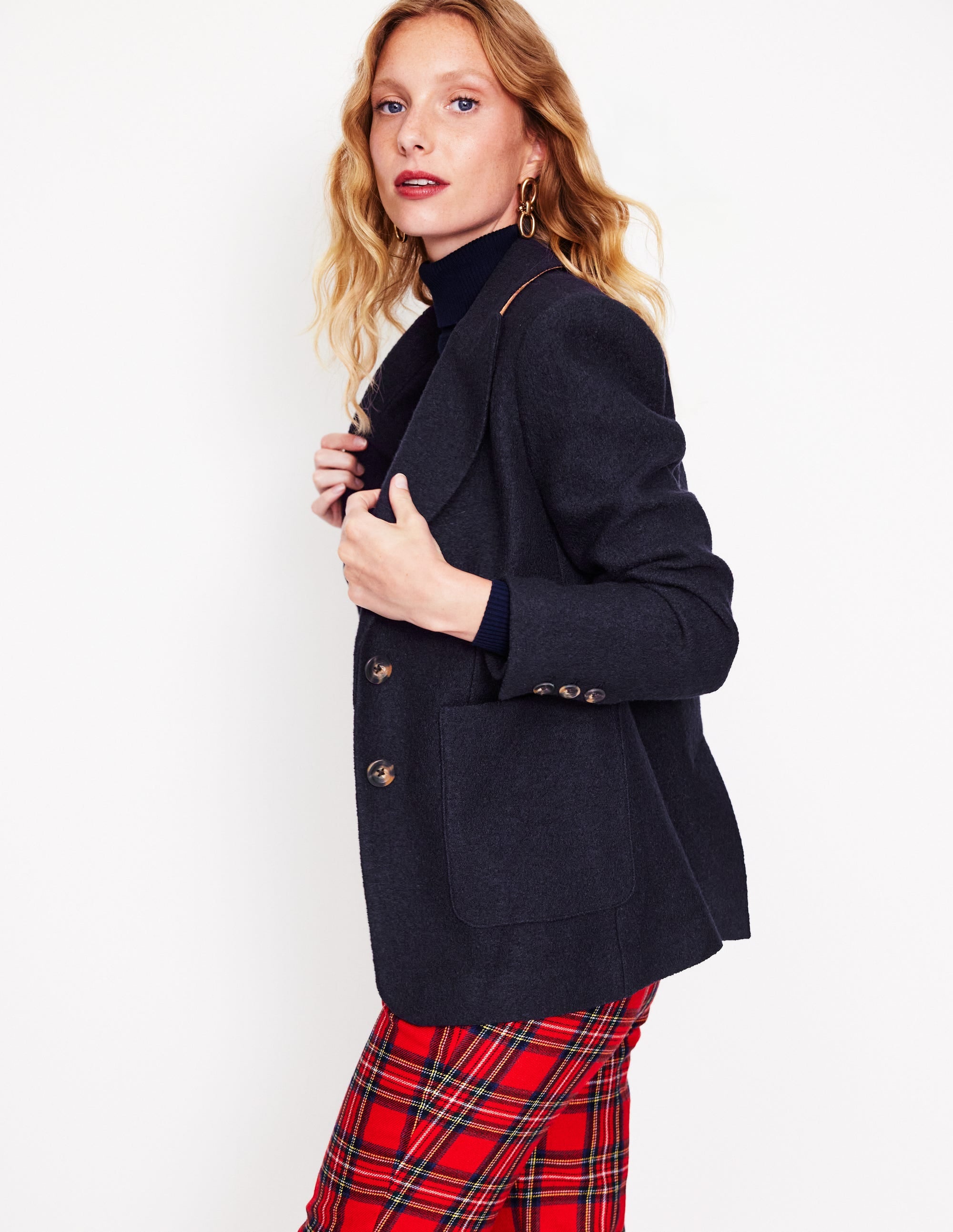 A woman wearing a navy textured blazer with a revere collar, contrast piping, and welt pockets, paired with red plaid pants.