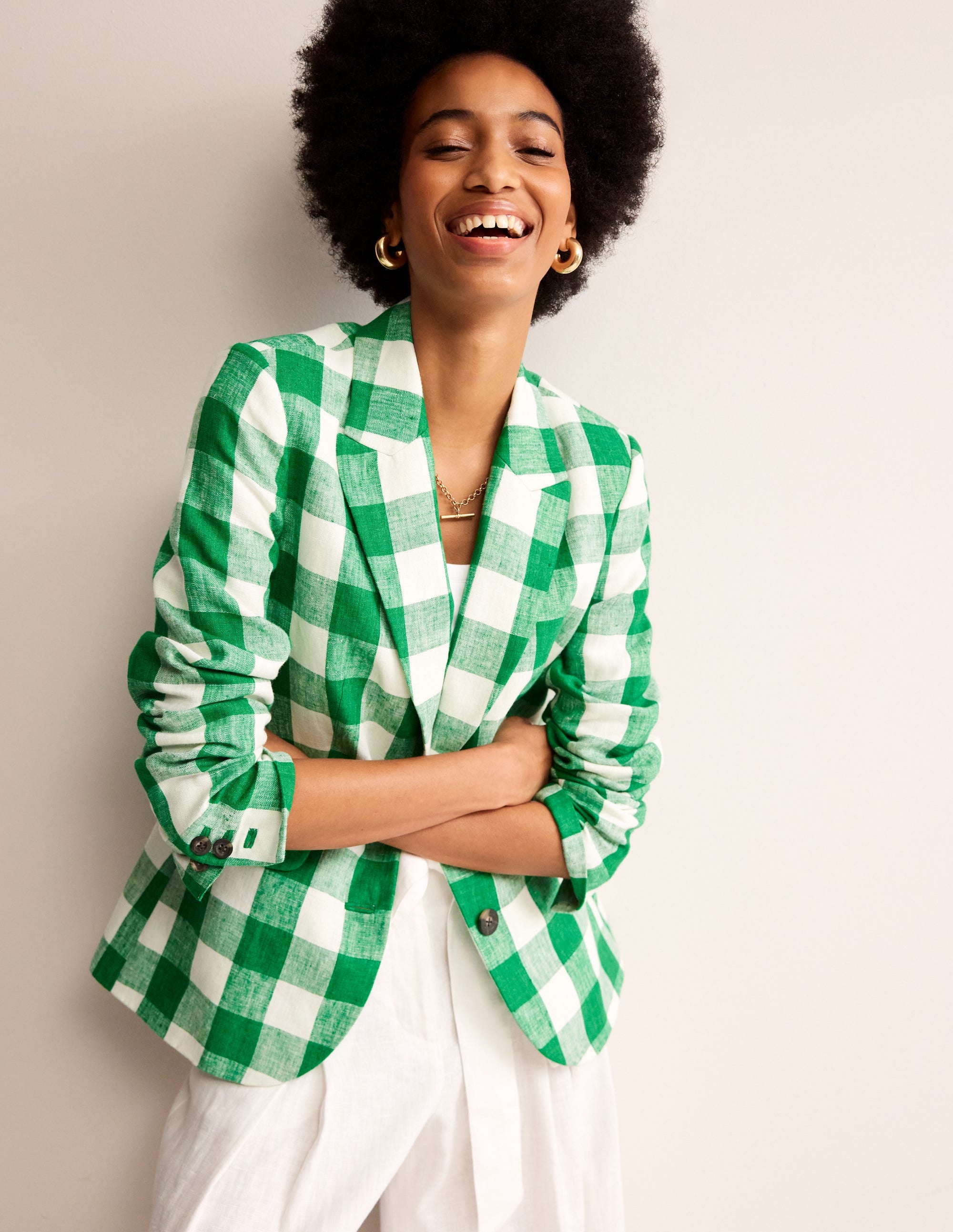 A woman wearing a green gingham linen blazer with a revere collar, contrast piping, and welt pockets, smiling with arms crossed.