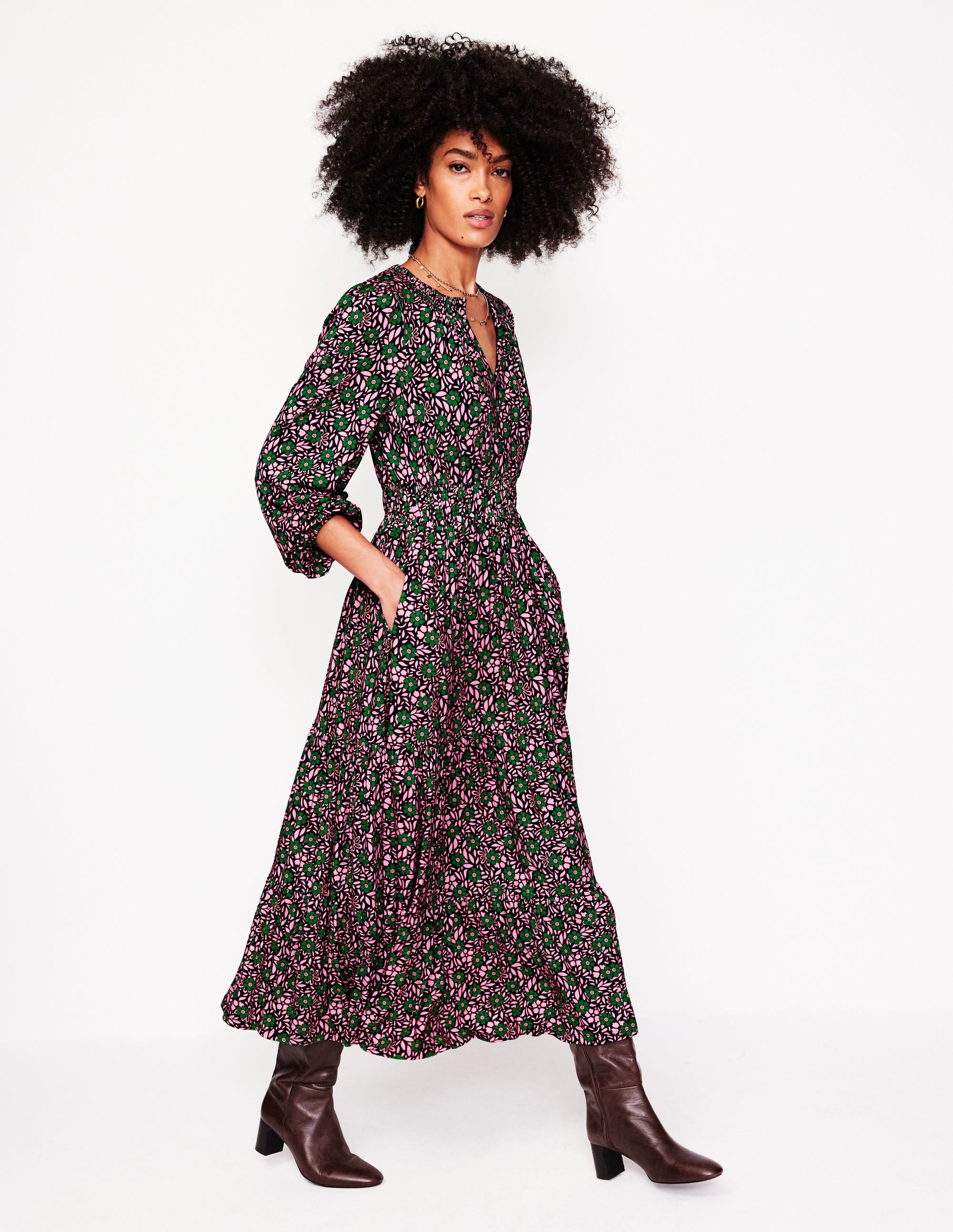 A woman wearing a long, floral print midi dress with long puffed sleeves, a fitted bodice, and a tiered skirt in sherbet pink, green, and black, paired with brown knee-high boots.