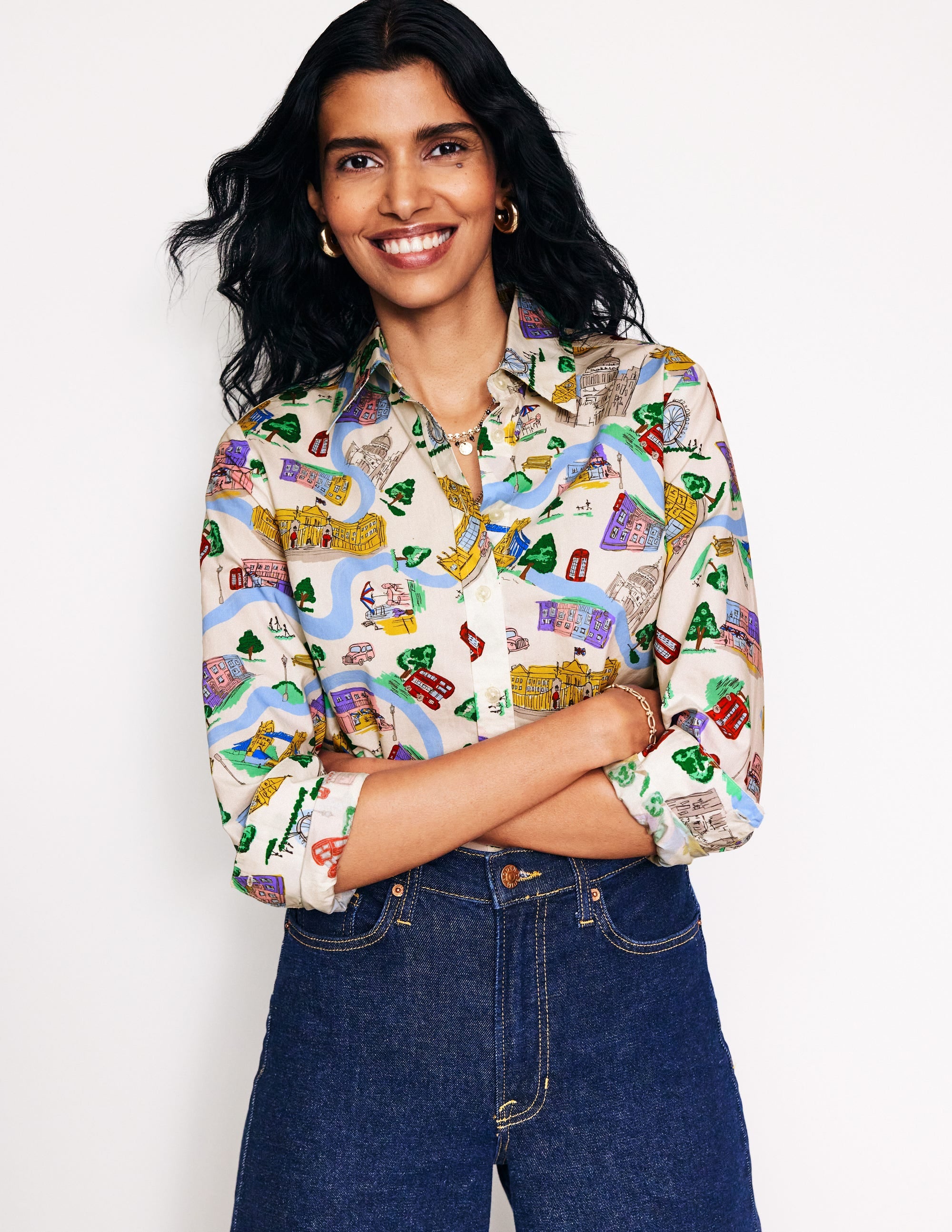 A woman wearing an ivory cotton shirt with a colorful London town print, long sleeves, and a classic collar, smiling with her arms crossed.