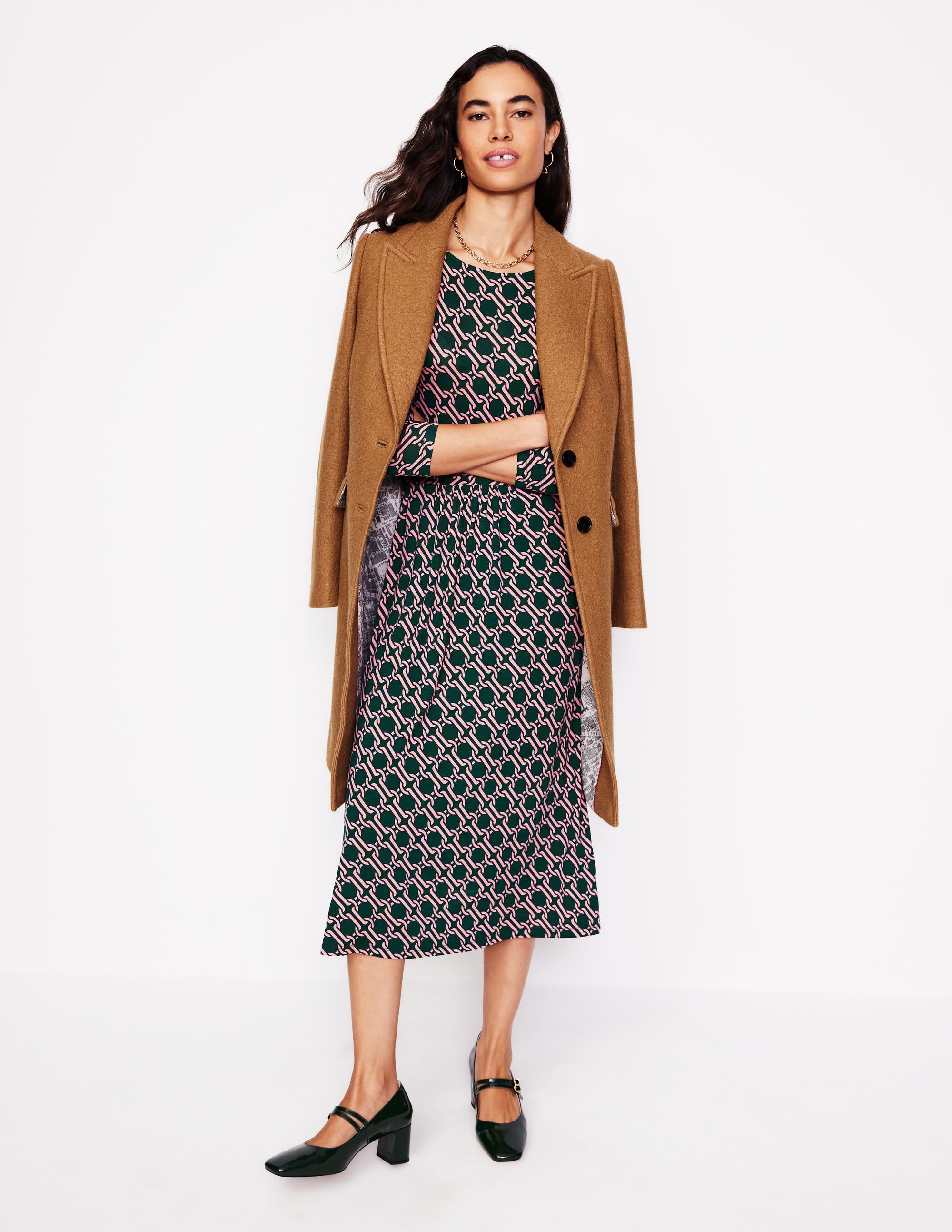 A woman wearing a midi dress with a pink and black chain geo pattern, long sleeves, and side seam pockets, paired with black heels and a brown coat.