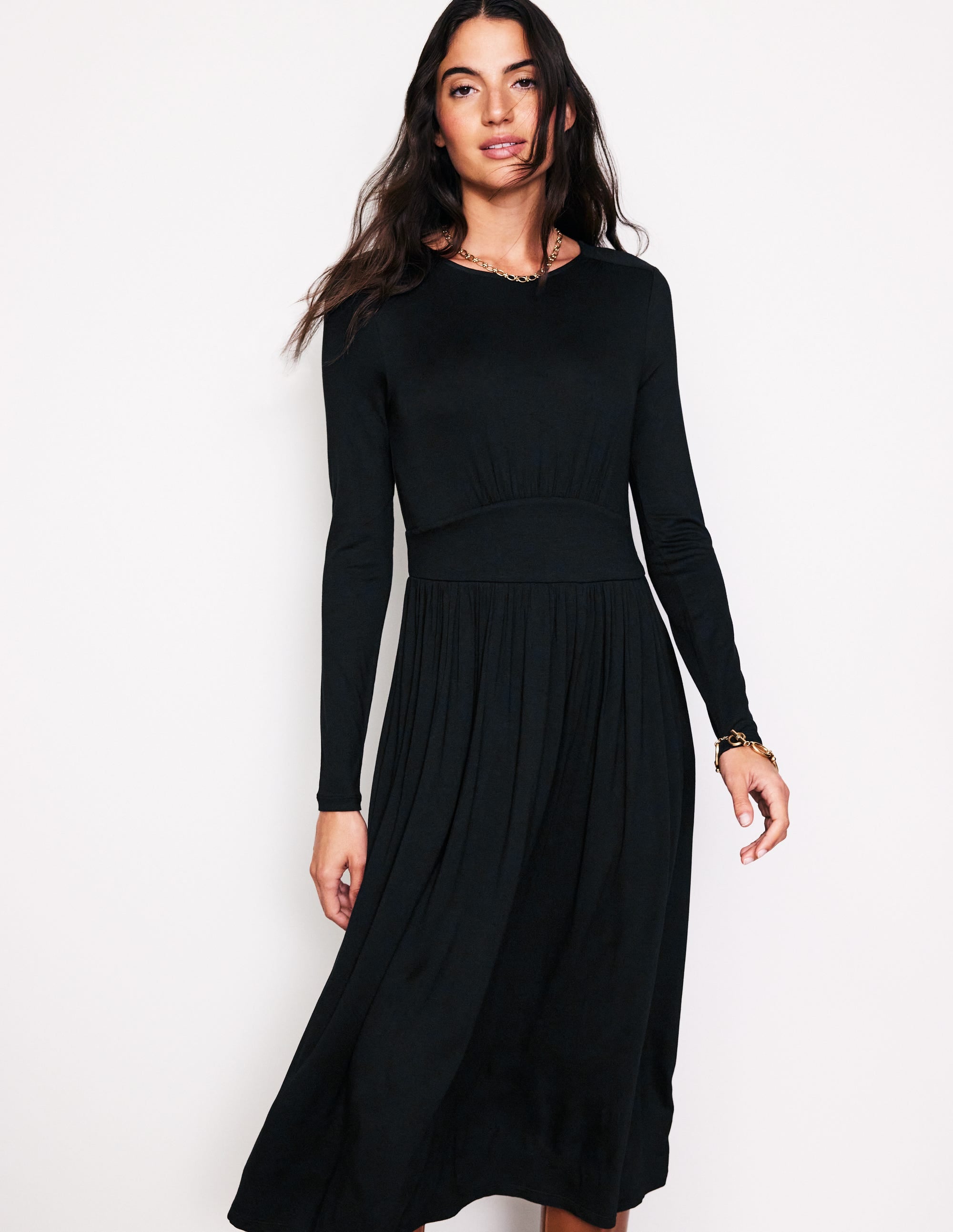 A woman wearing a black long sleeve jersey dress with a gathered skirt and waist panel, standing against a plain background.