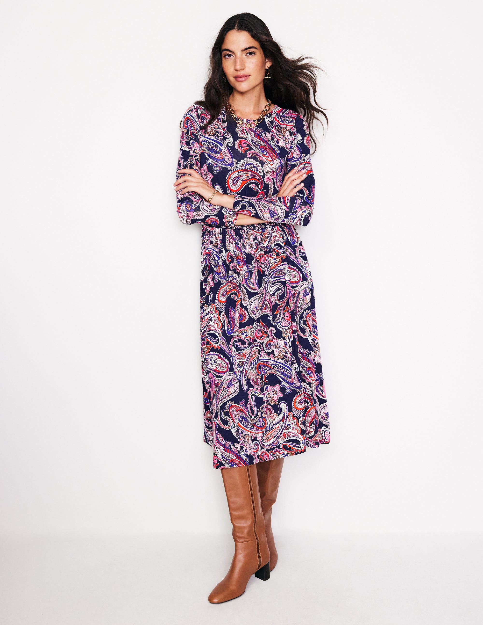 A woman wearing a long sleeve dress with a colorful paisley pattern, gathered skirt, and waist panel, paired with brown knee-high boots.