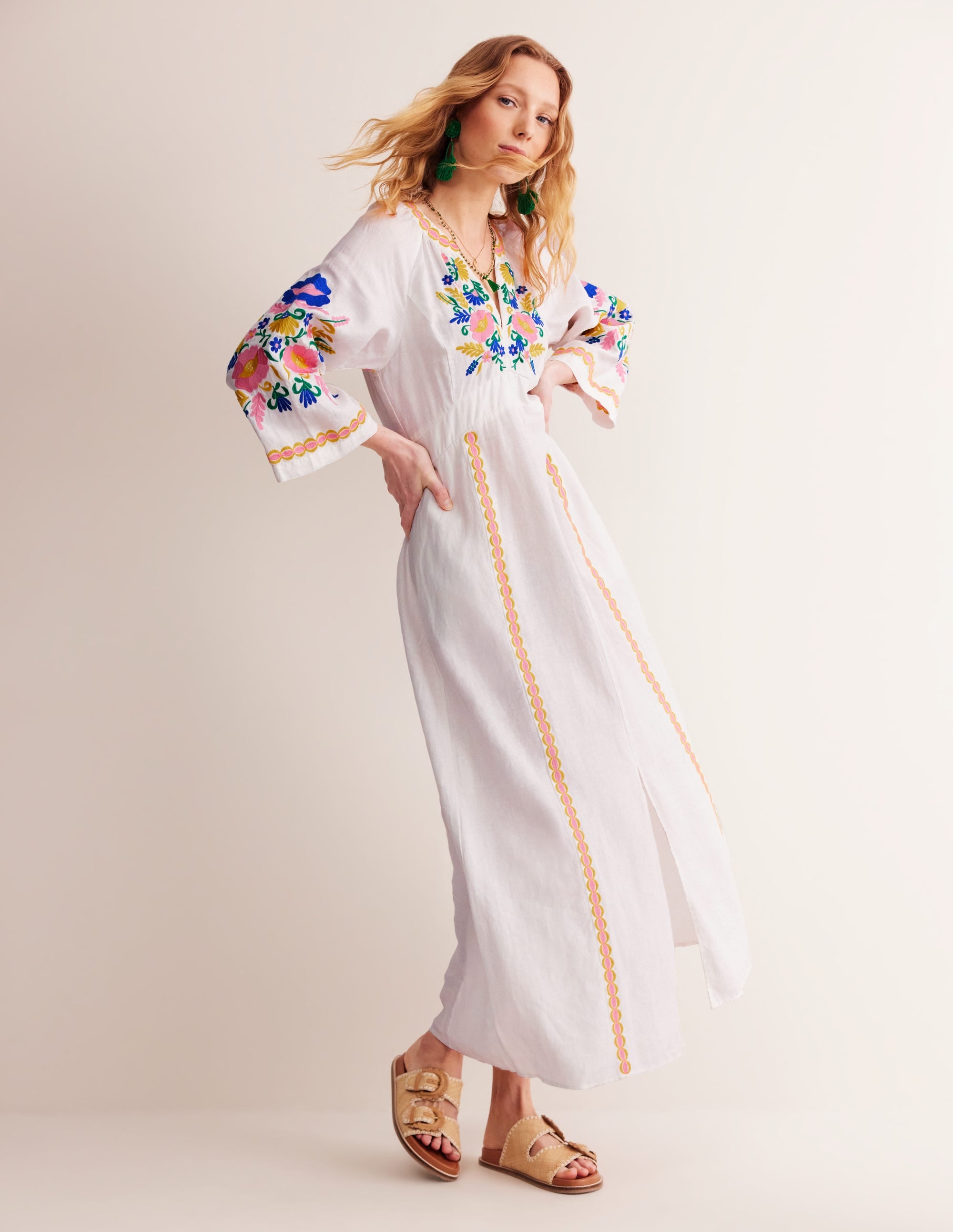 A woman wearing a long, ivory linen embroidered dress with colorful floral embroidery on the bodice and sleeves, featuring side seam pockets, and styled with green earrings and beige sandals.