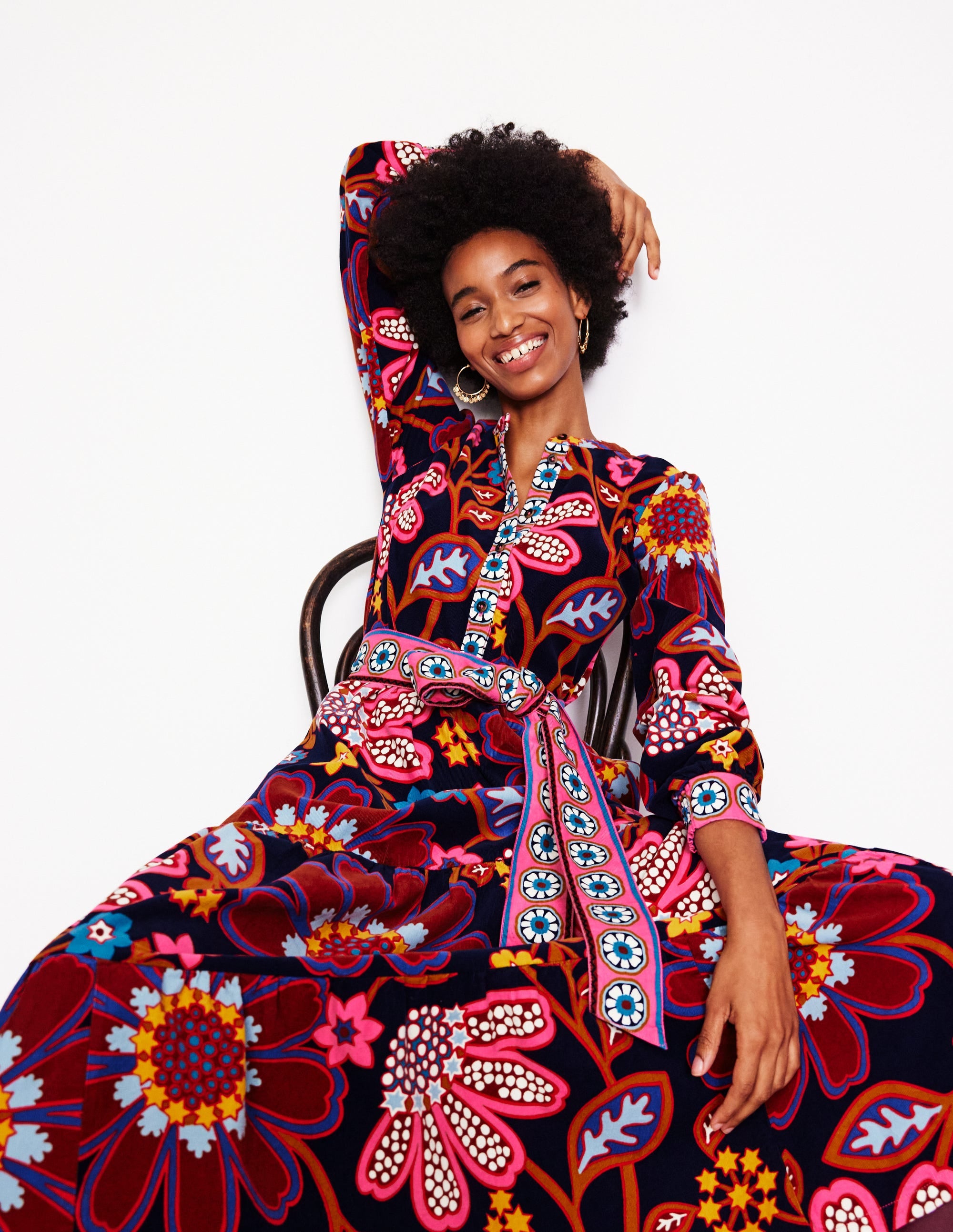 A woman wearing a colorful, patterned maxi dress with long sleeves, a tiered design, and a tie waist, sitting on a chair and smiling.
