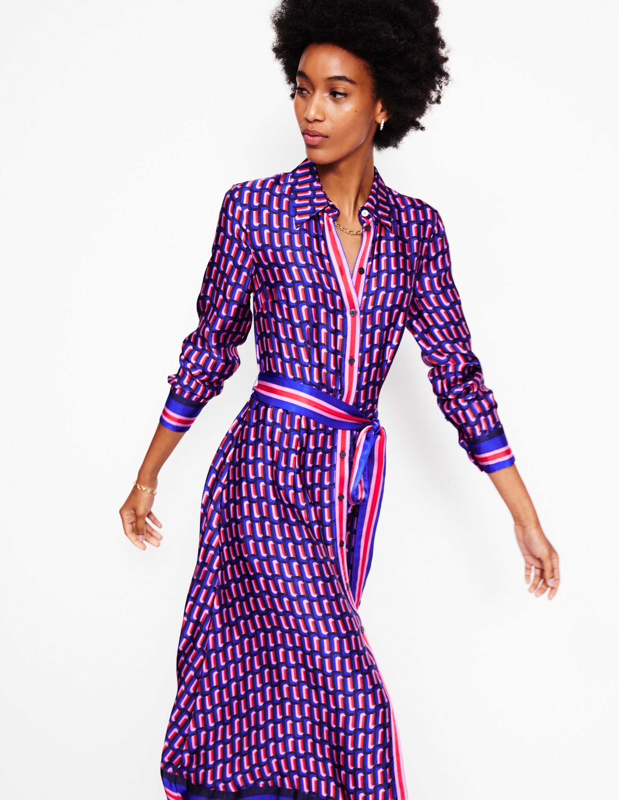 A woman wearing a Mazarine Blue satin shirt dress with a geometric pattern, long sleeves, a collar, and a belt, standing against a plain background.