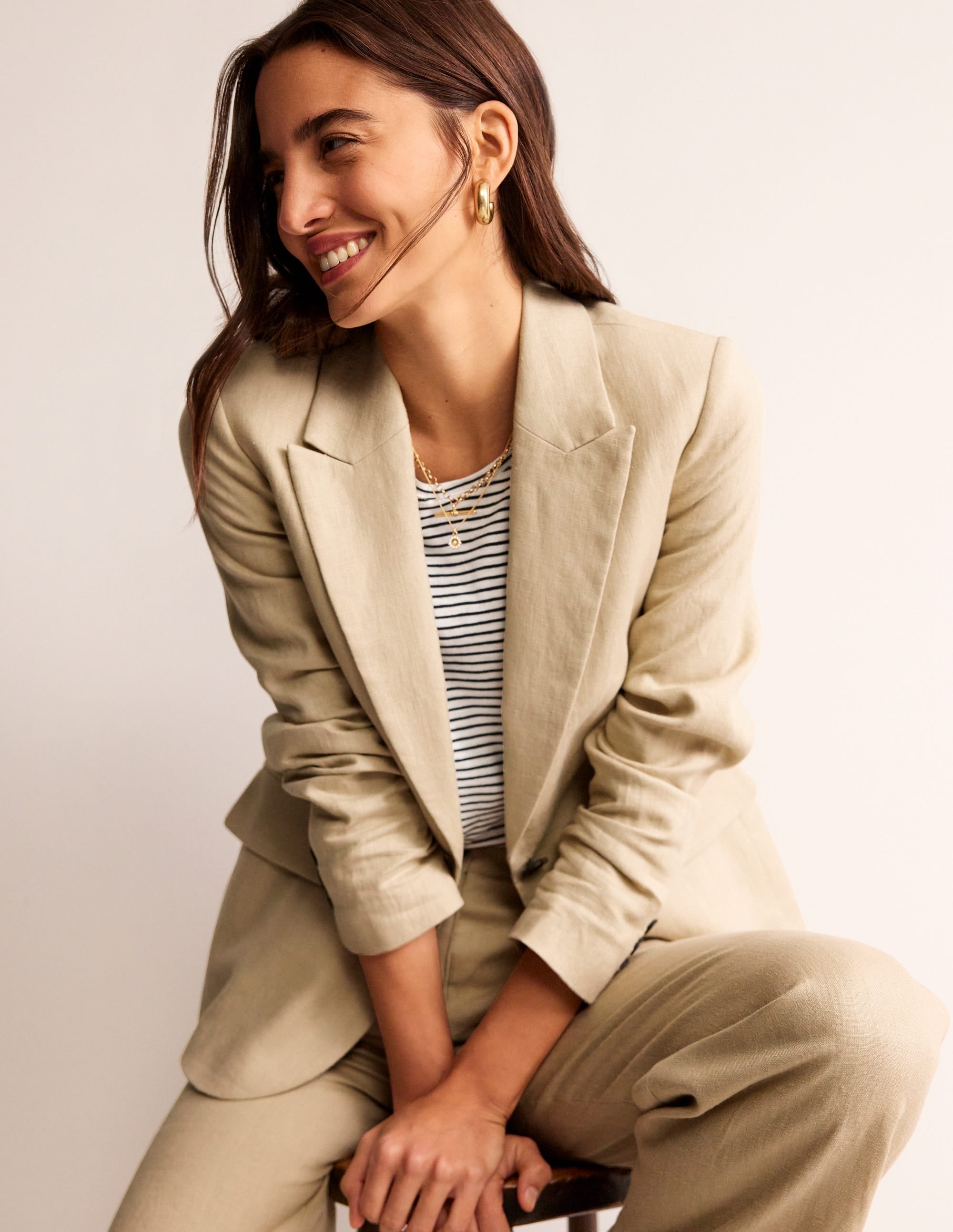A woman wearing a beige linen blazer with a revere collar, contrast piping, and welt pockets, paired with a striped top and gold jewelry.