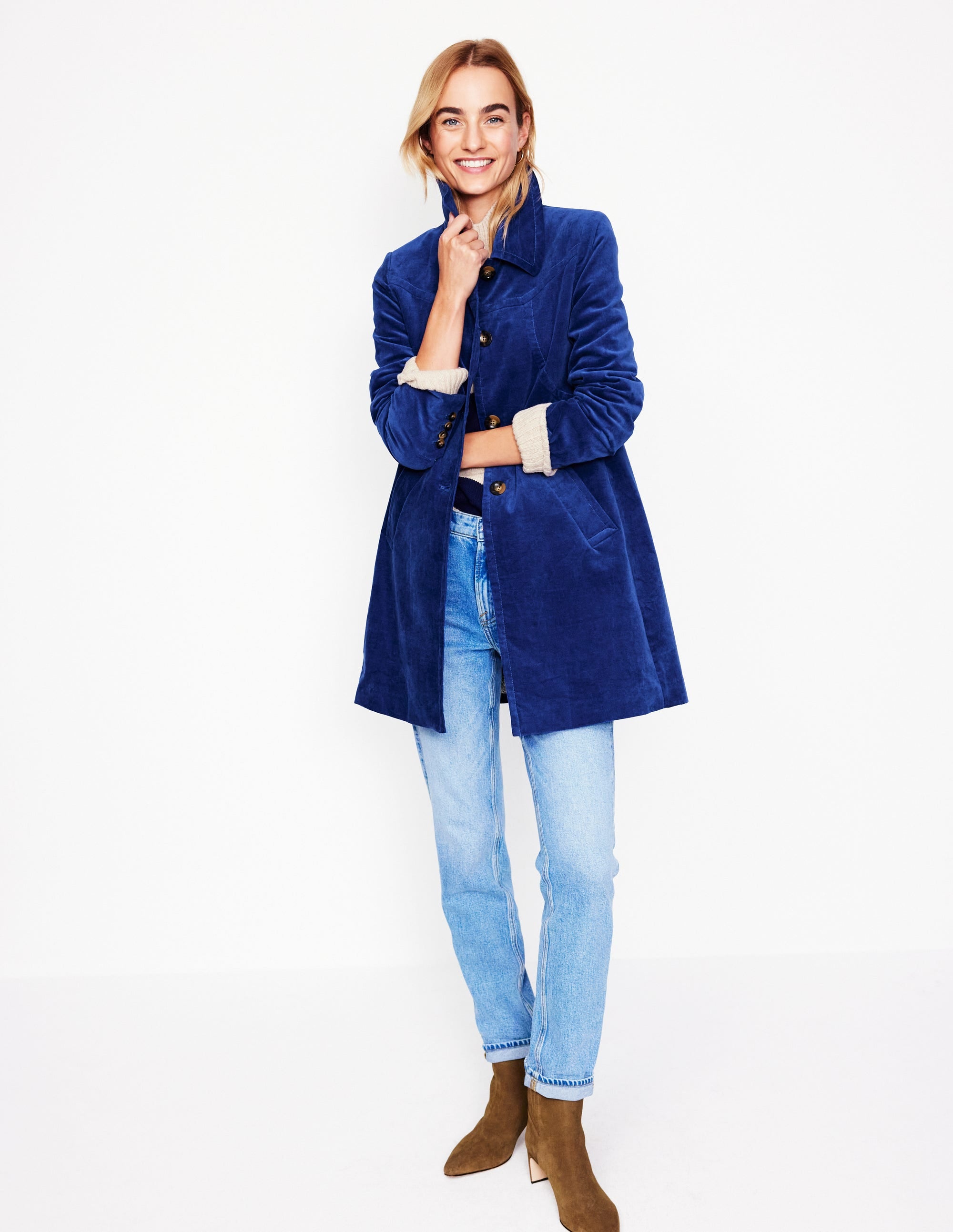 A woman wearing a blue velvet coat with a classic collar, long sleeves, and a straight fit, paired with light blue jeans and brown ankle boots.