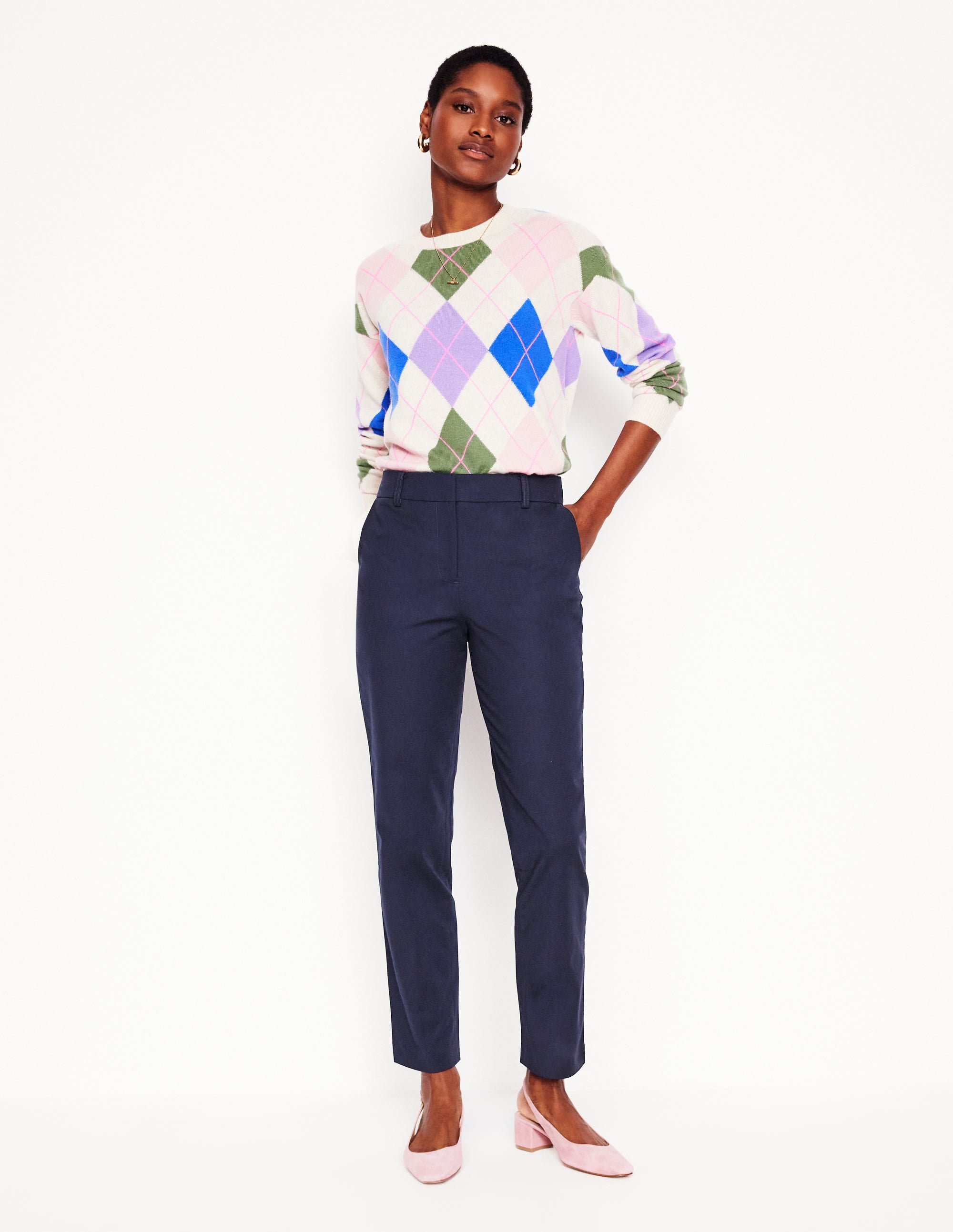 A woman wearing navy Richmond 7/8 trousers with a fitted waist, functional pockets, and a tapered, relaxed fit above the ankle, paired with a colorful argyle sweater.