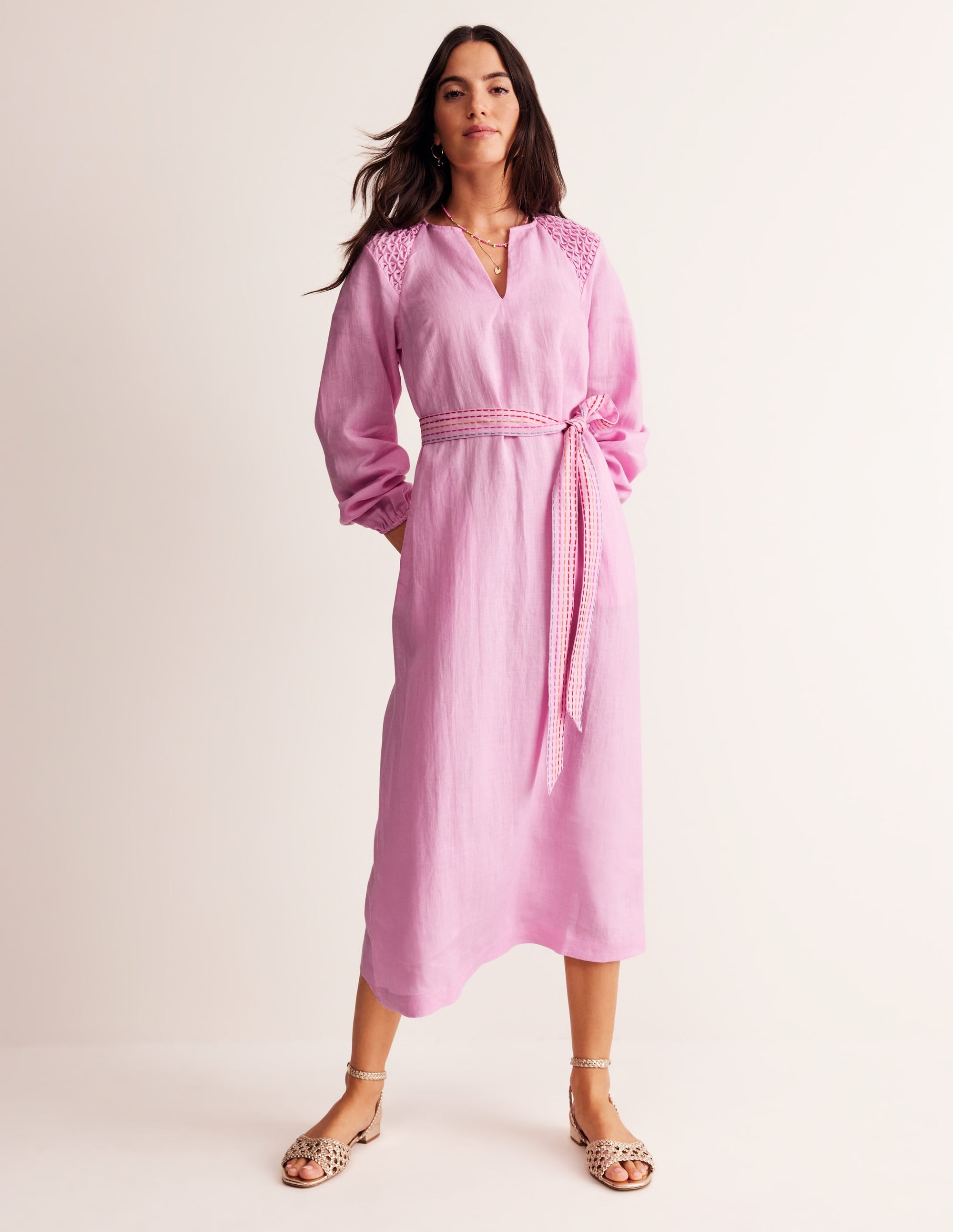 A woman wearing a sweet lilac linen midi dress with blouson sleeves, smocking on the shoulders, a self-fabric belt, and side pockets.
