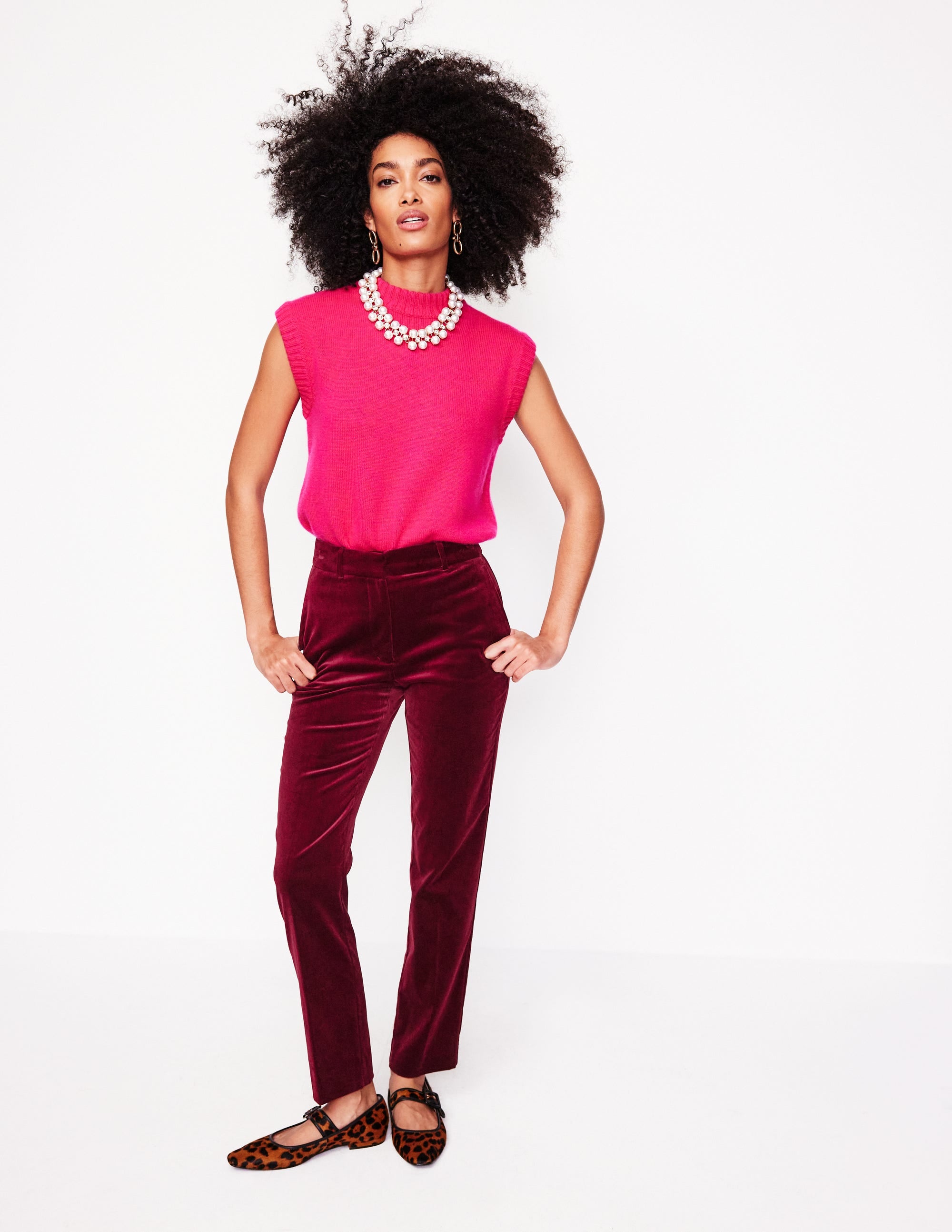 A woman wearing deep red velvet trousers with a tapered leg, paired with a bright pink sleeveless top, accessorized with a pearl necklace and earrings.