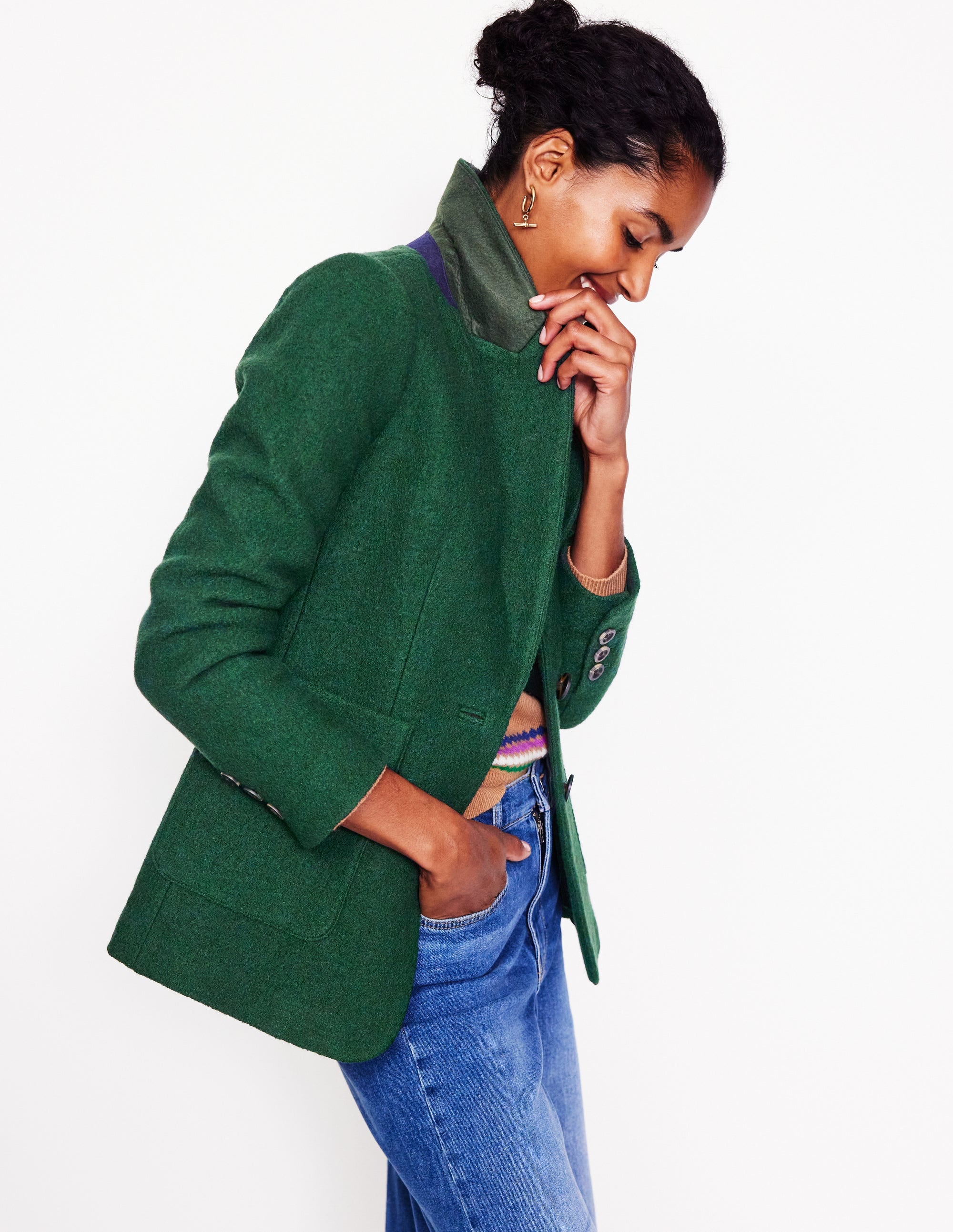 A woman wearing a dark green textured blazer with a revere collar, contrast piping, and welt pockets.