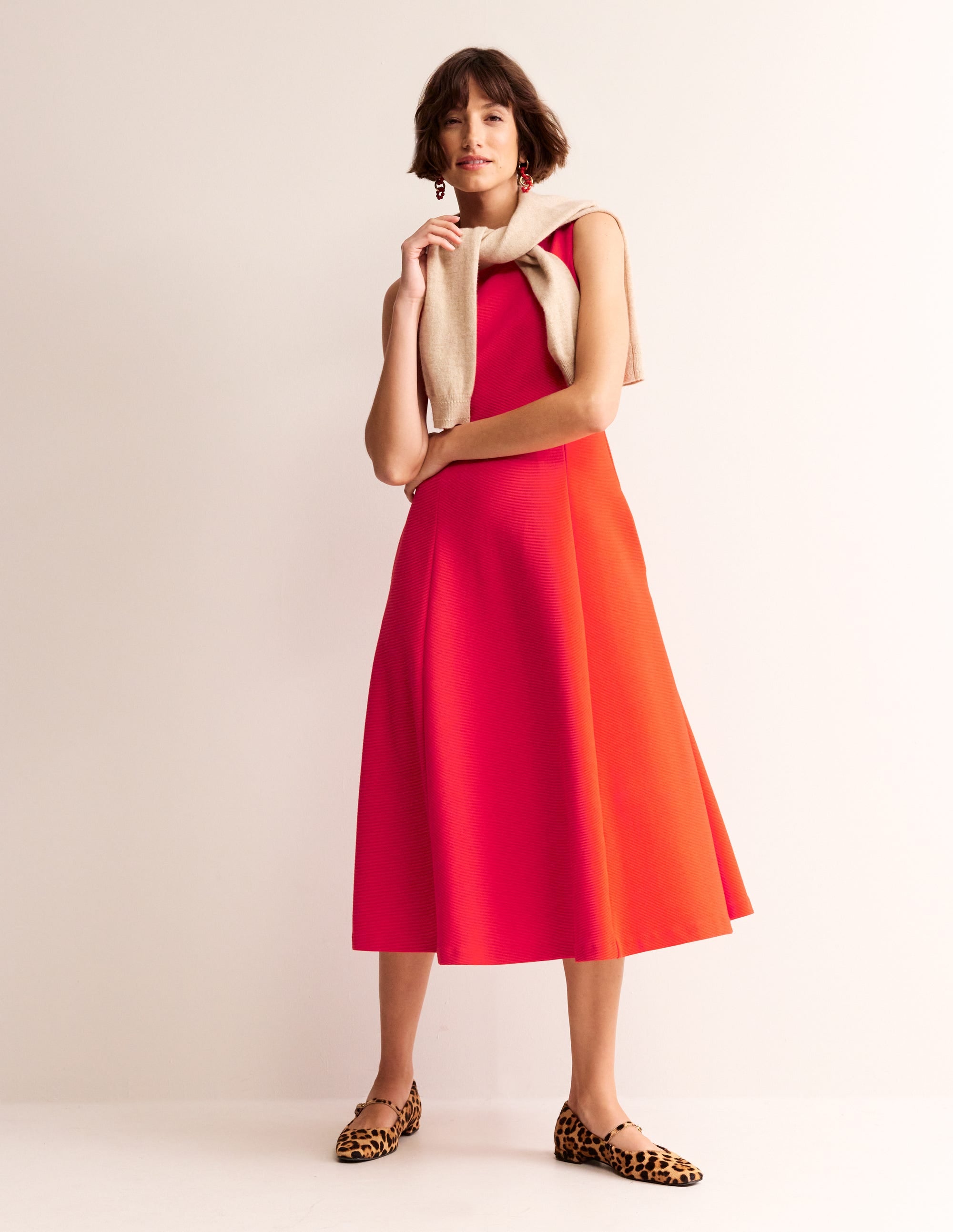 A woman wearing a flame scarlet ponte dress with a fitted bodice and flared skirt, paired with leopard print shoes and a beige sweater draped over her shoulders.