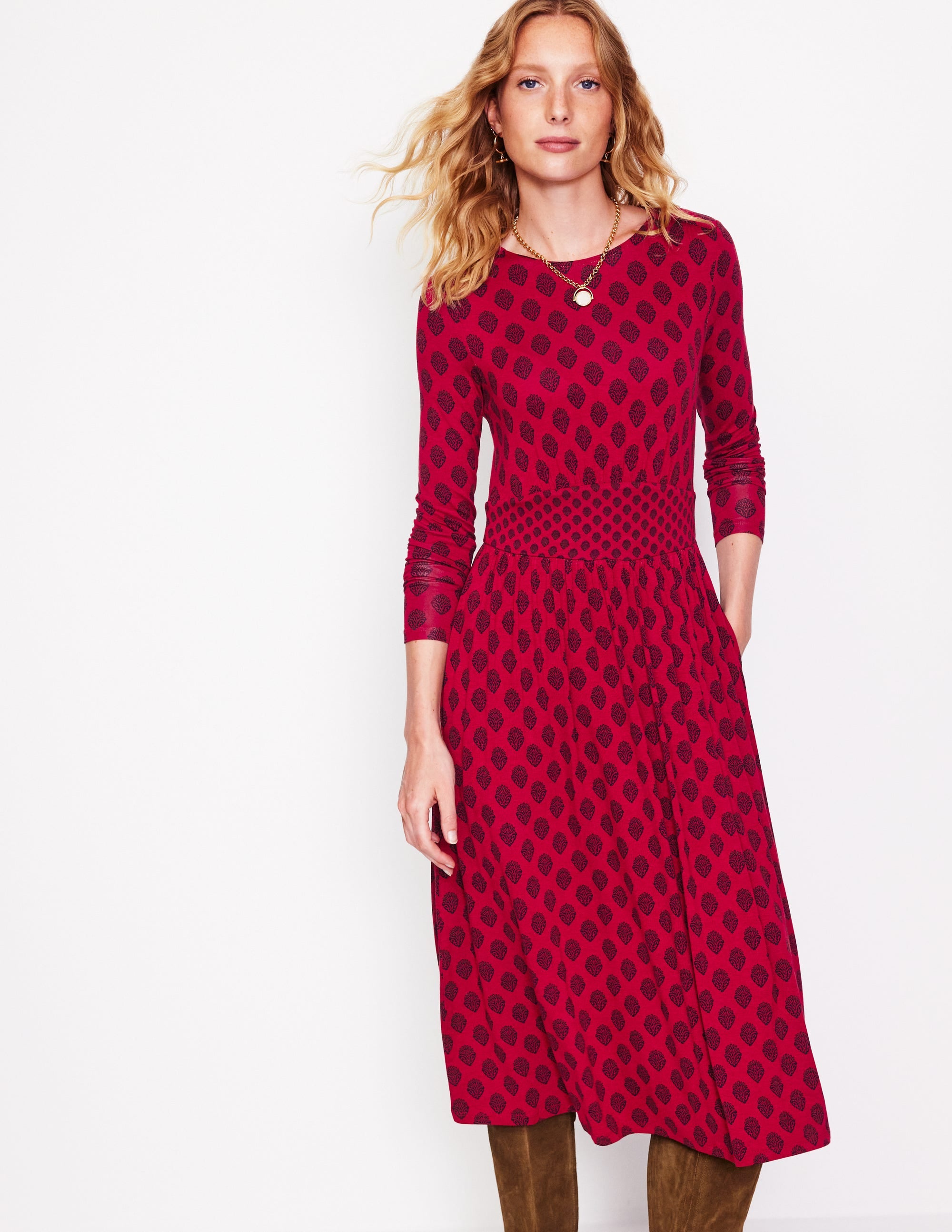 A woman wearing a long sleeve red jersey dress with a black ornate stamp pattern, fitted waist, and gathered skirt.