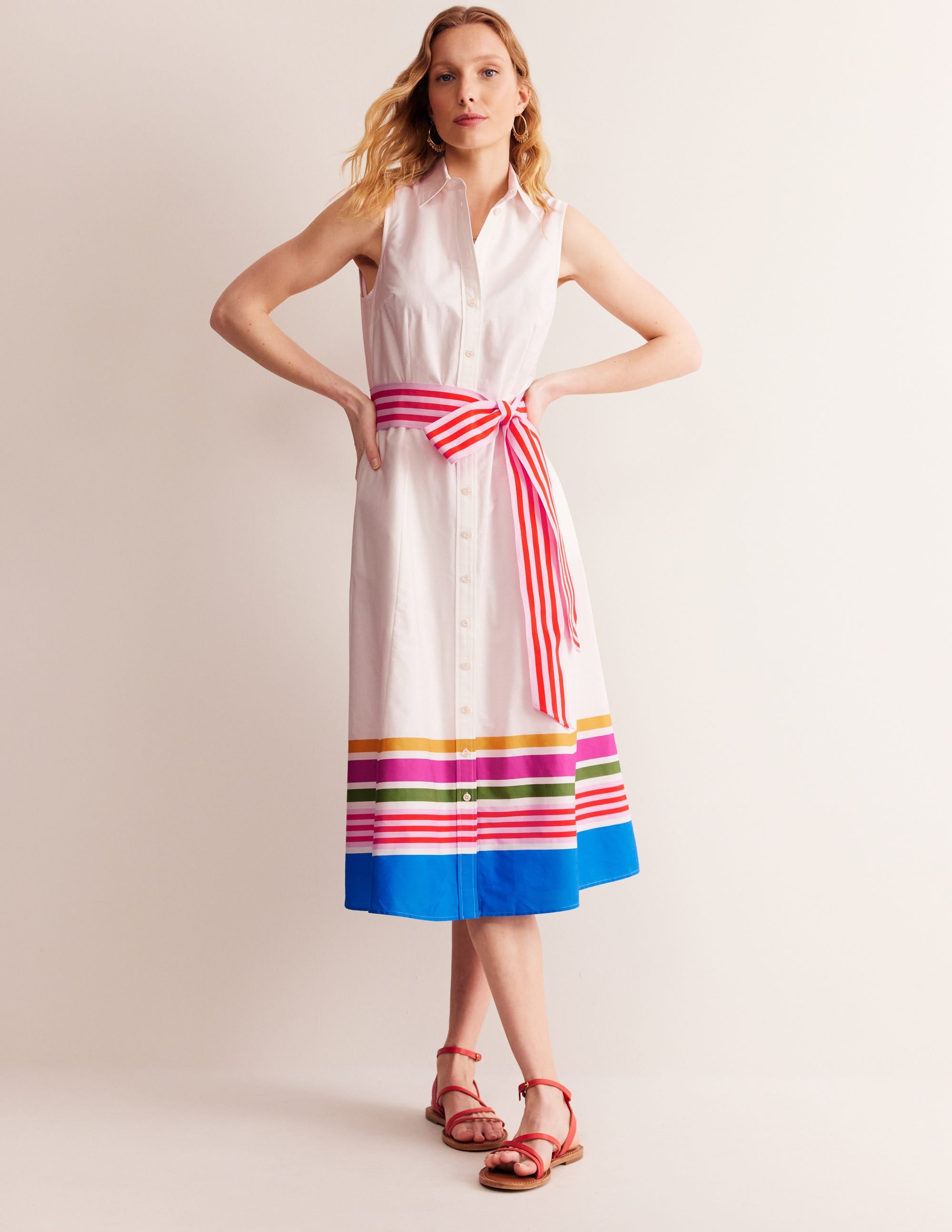 A woman wearing a sleeveless ivory shirt dress with multicolored horizontal stripes at the hem, a belted waist with a striped sash, and a classic collar.