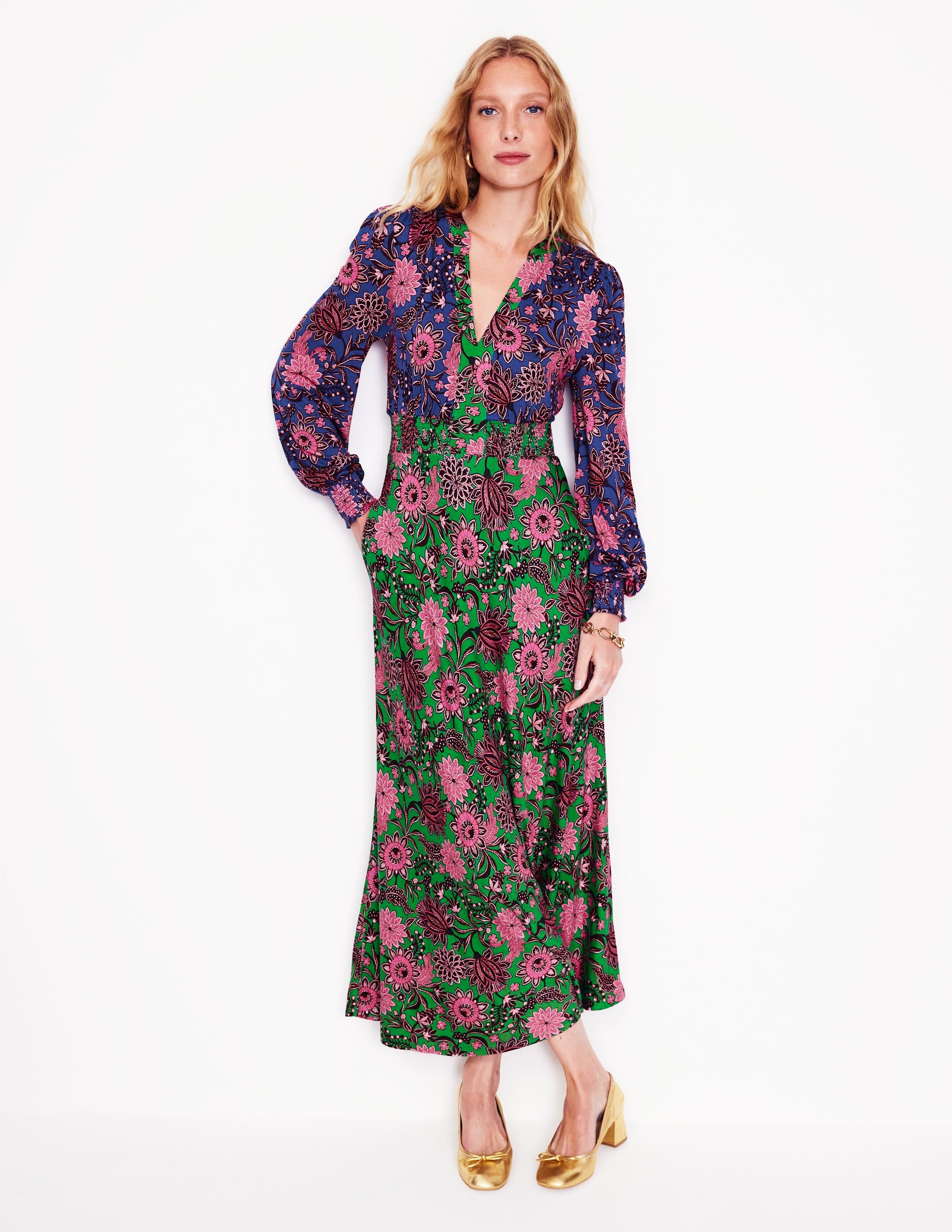 A woman wearing a long, floral midi dress with a smocked waist, notch neckline, and long sleeves, in rich emerald green with pink and purple floral patterns.