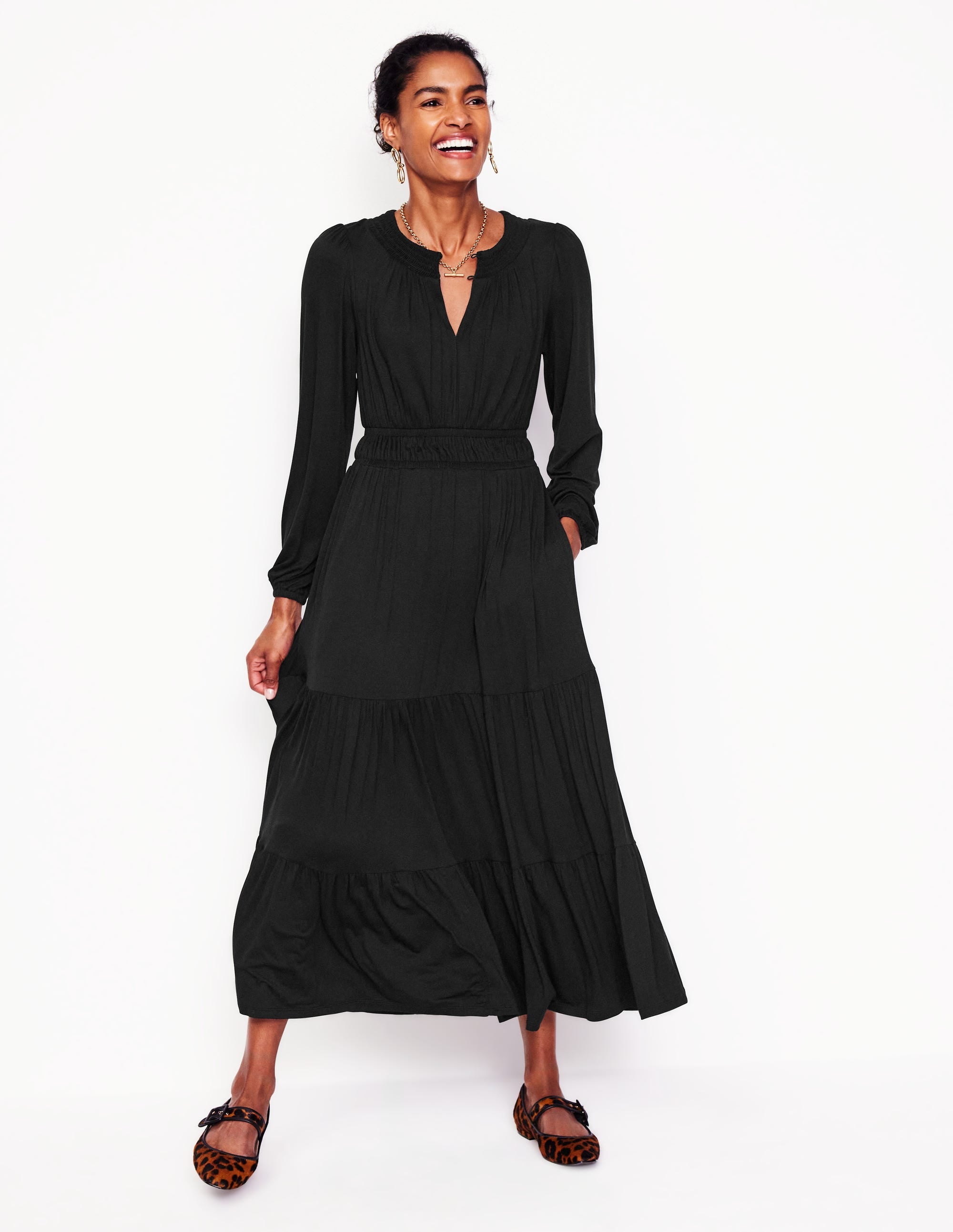 A woman wearing a black smock tiered jersey midi dress with long sleeves and pockets, standing against a plain background.
