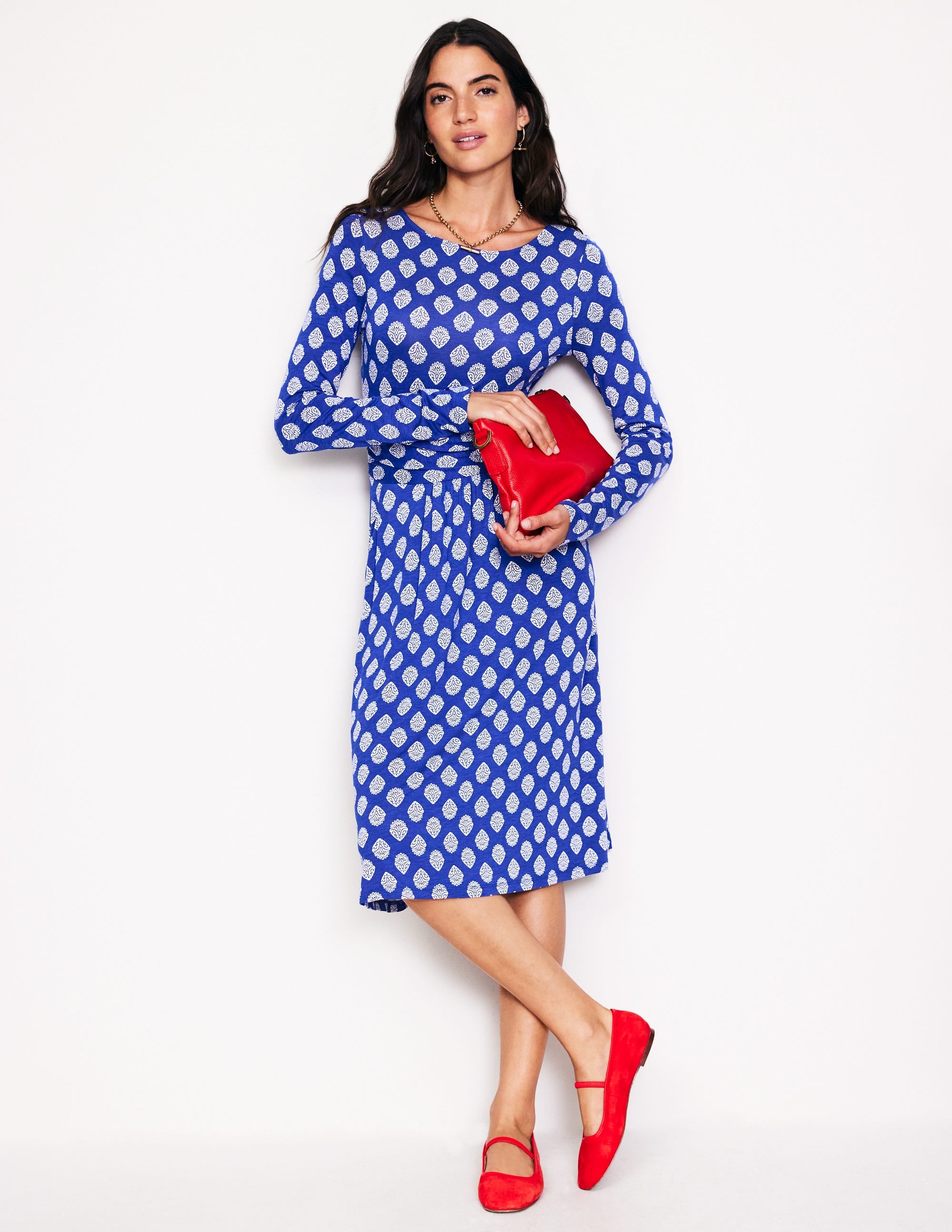 A woman wearing a blue long sleeve dress with a white ornate stamp pattern, holding a red clutch, and wearing red shoes.
