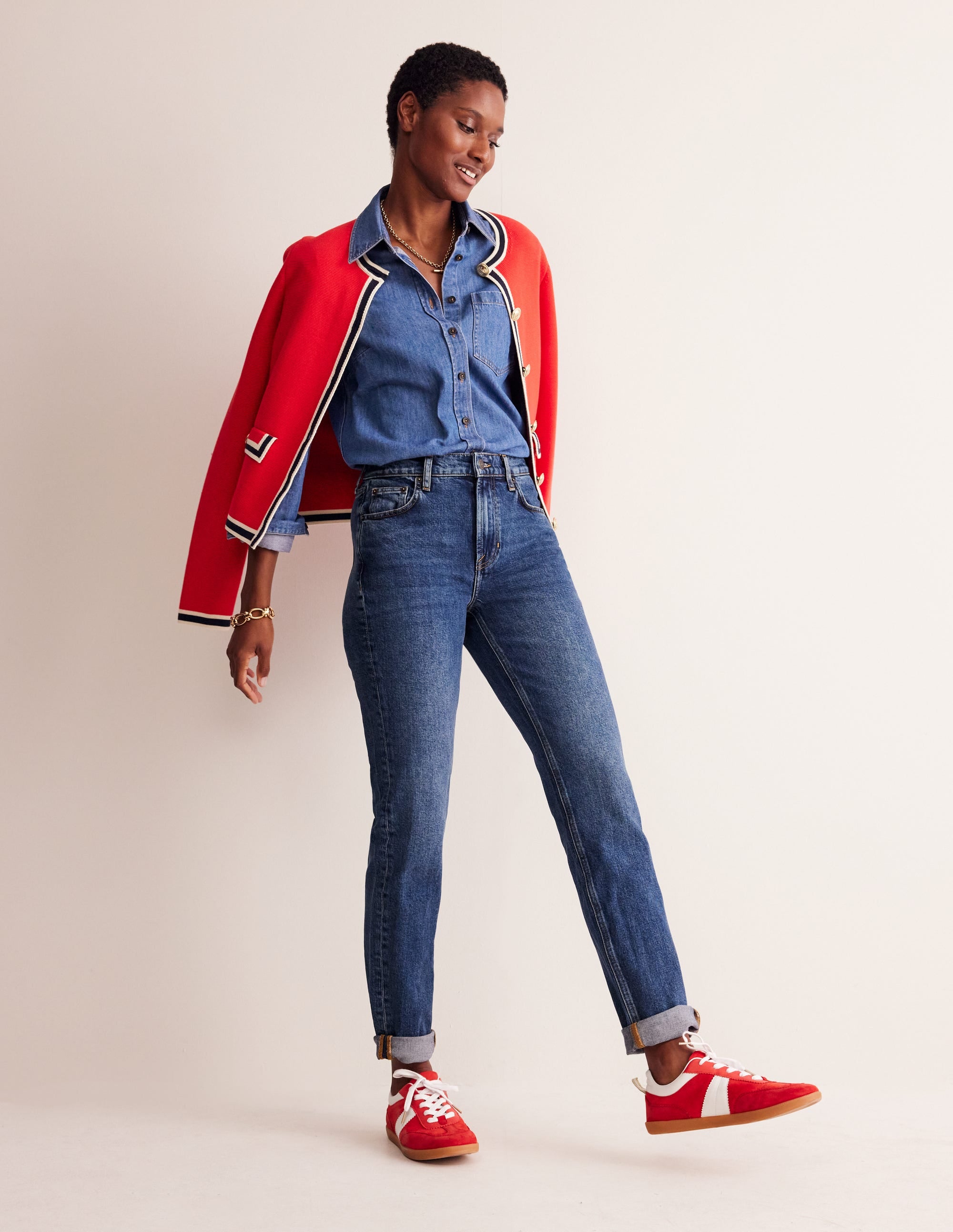 A woman wearing blue relaxed-fit jeans with rolled-up hems, a blue denim shirt, red sneakers, and a red jacket draped over her shoulders.
