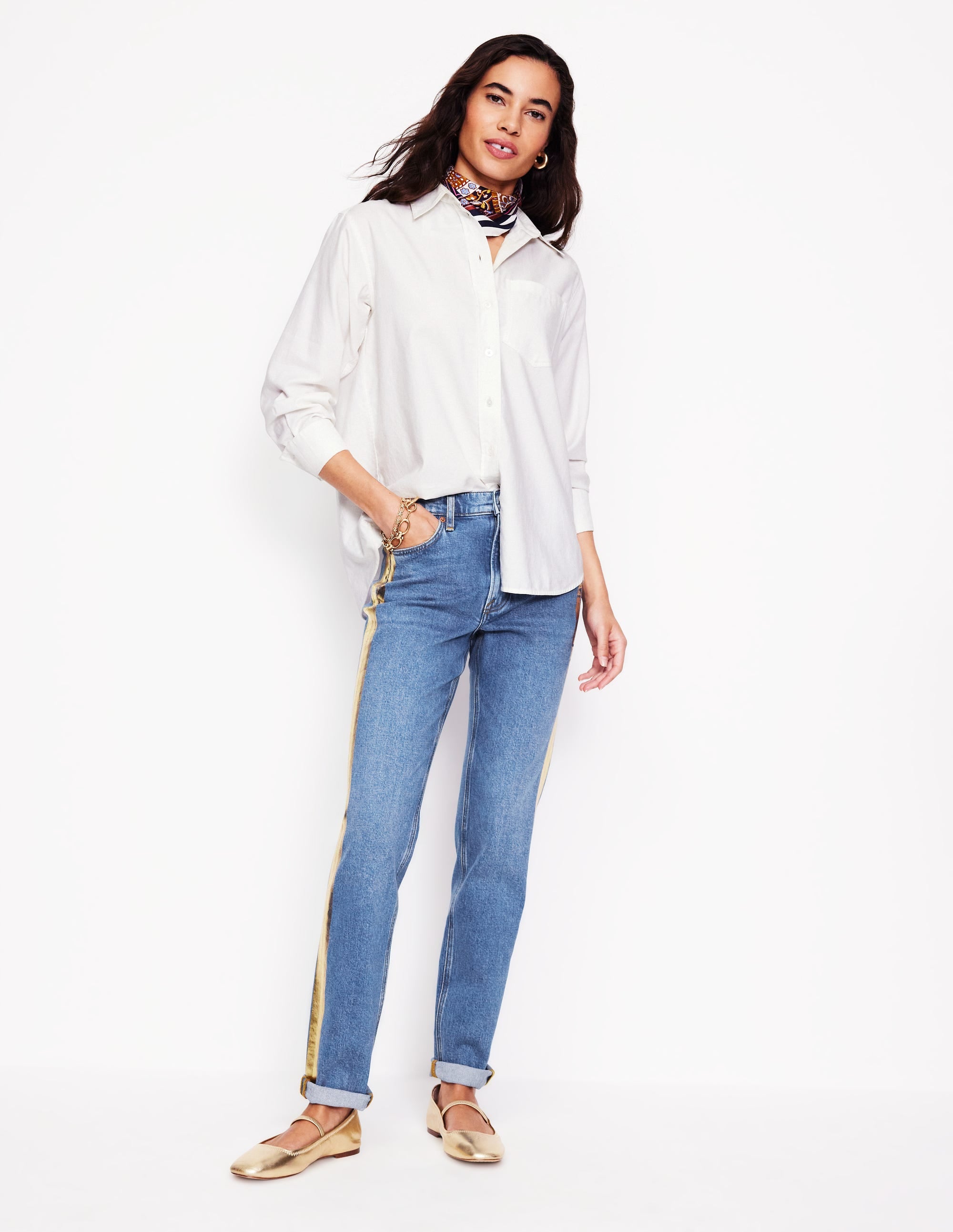 A woman wearing medium blue girlfriend jeans with a metallic gold side stripe and rolled-up hems, paired with a white shirt and gold shoes.