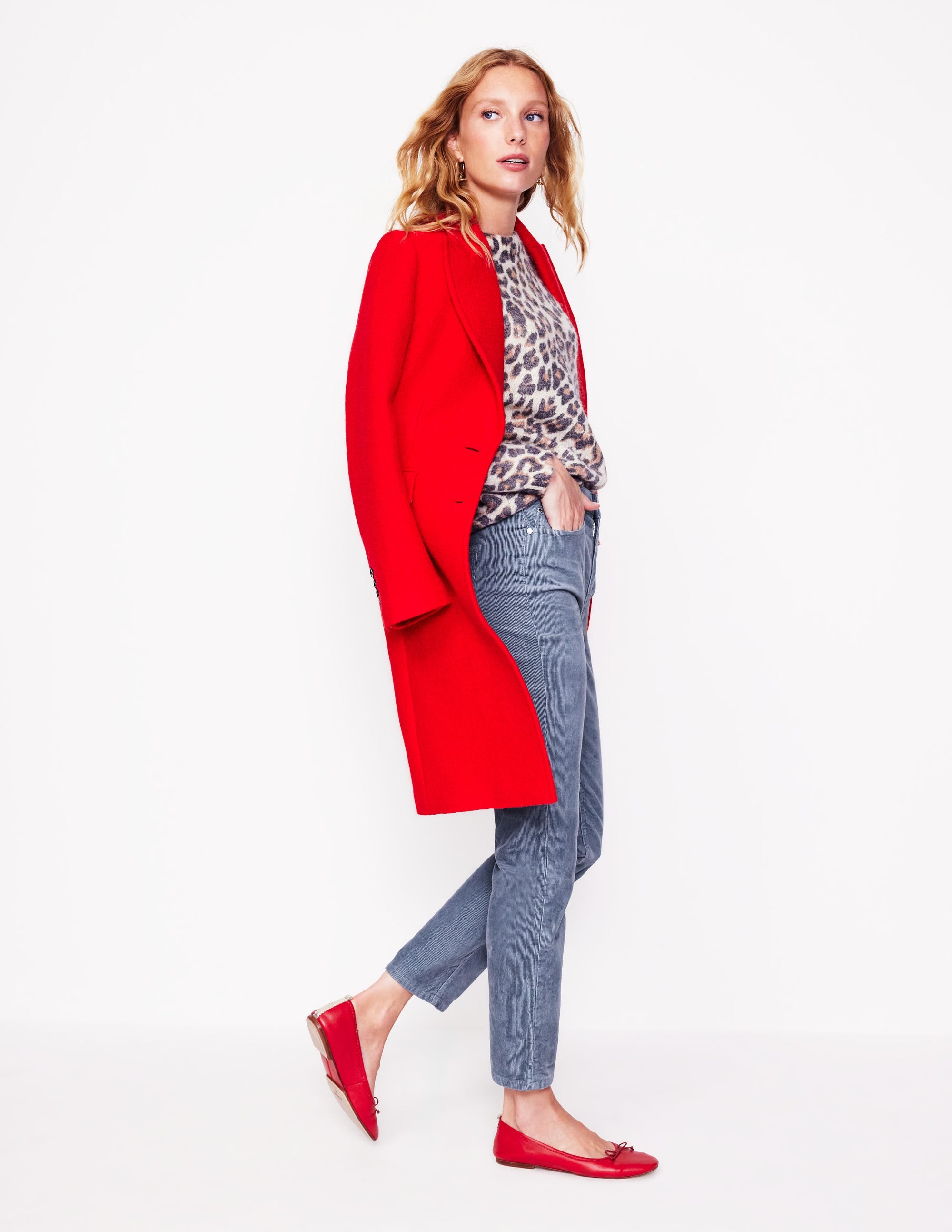 A woman wearing grey slim-fit corduroy jeans with five pockets, a button and zip fly, paired with a leopard print top, red coat, and red flats.