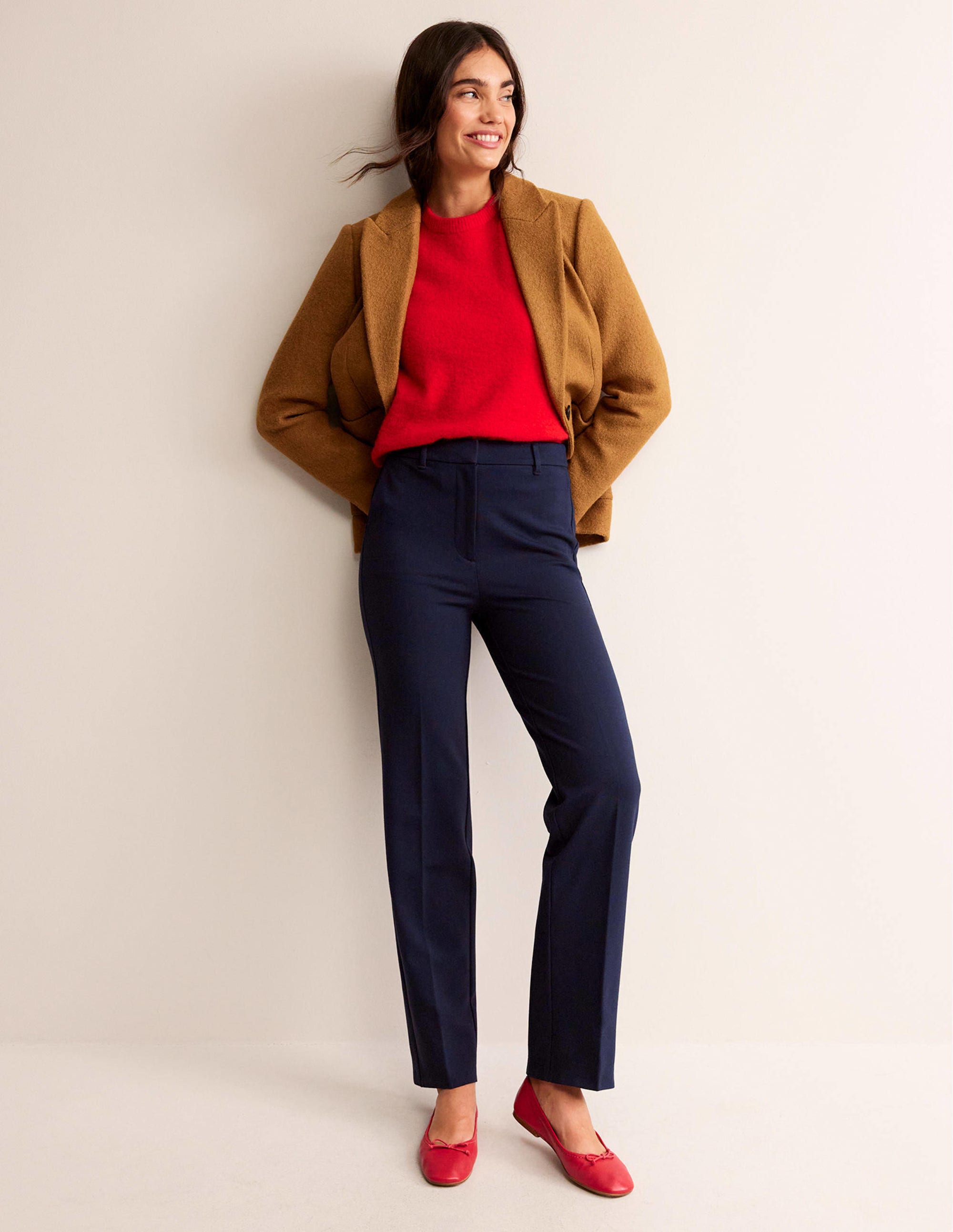 A woman wearing navy straight-leg trousers, a red top, a brown jacket, and red shoes, standing against a plain background.