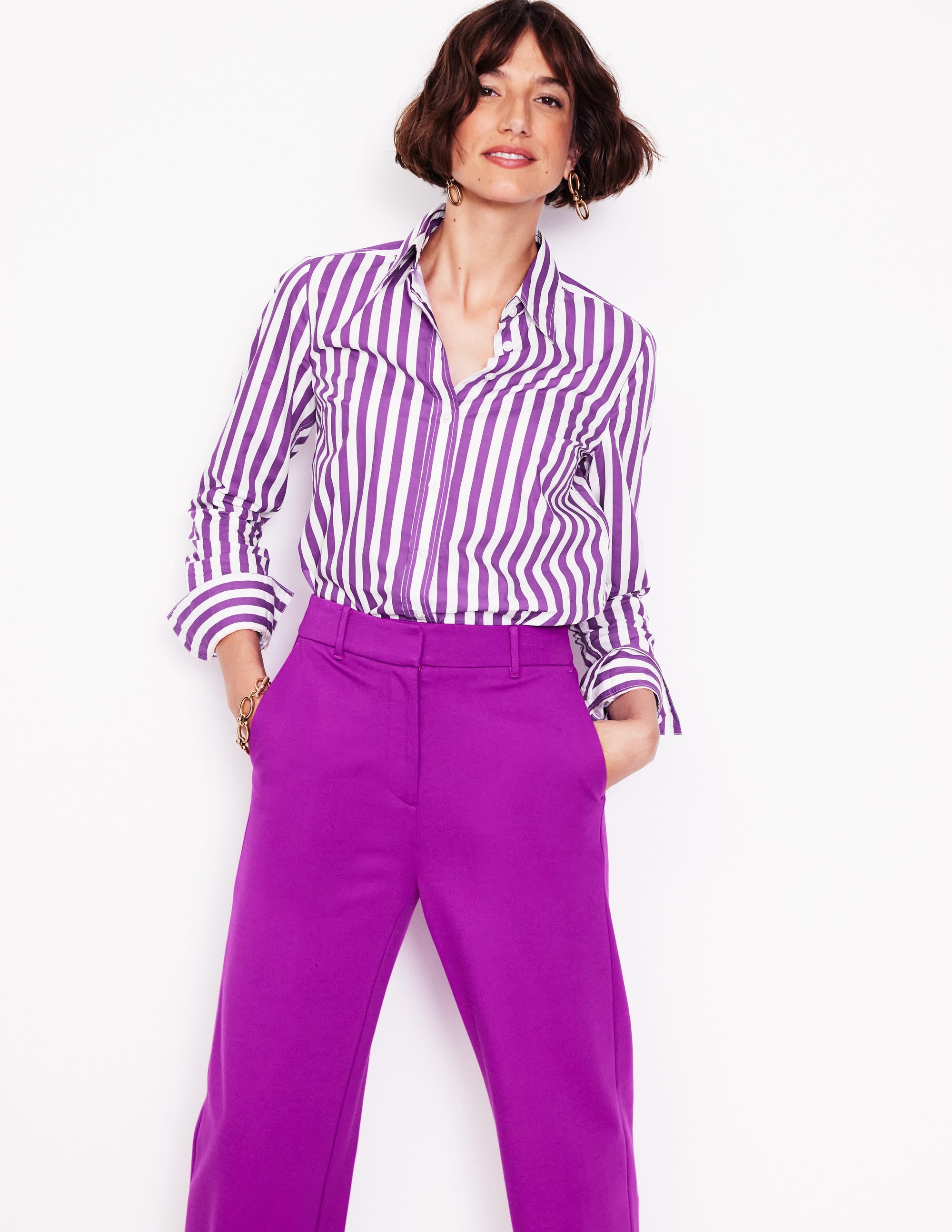 A woman wearing a royal purple and ivory striped cotton shirt with a classic collar and curved hem, styled with purple pants.