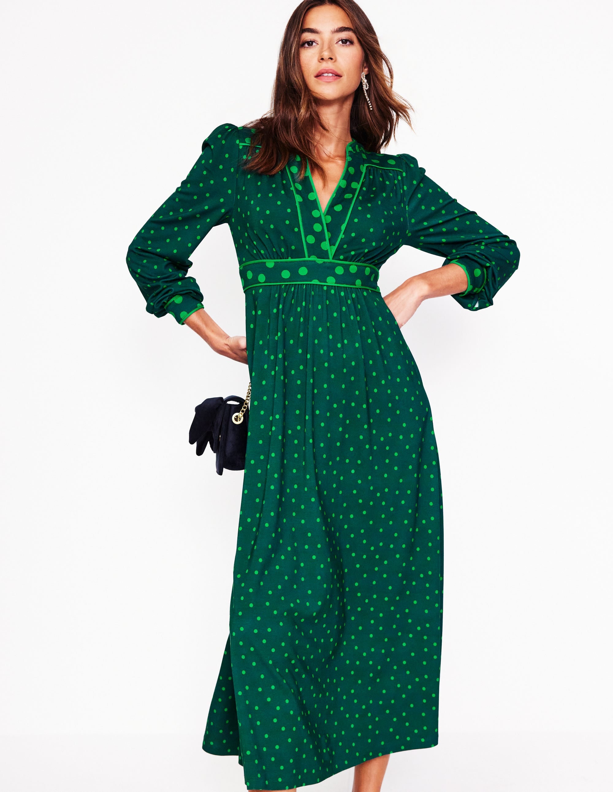 A woman wearing a green structured dress with a hotchpotch dot pattern, contrast piping, and side seam pockets.
