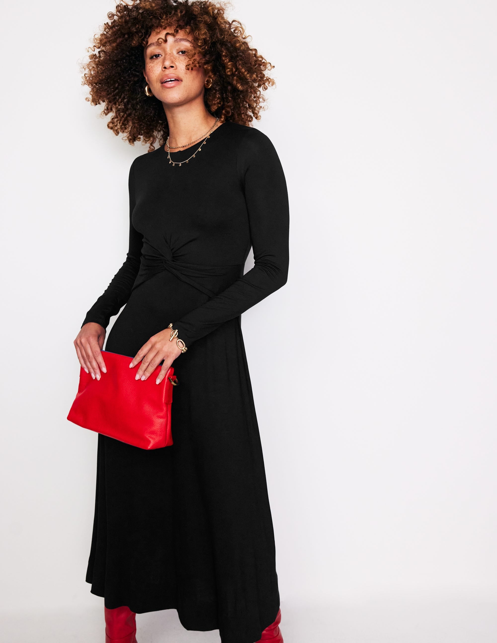 A woman wearing a black twisted waist jersey dress with long sleeves and a fitted bodice, standing against a plain background.