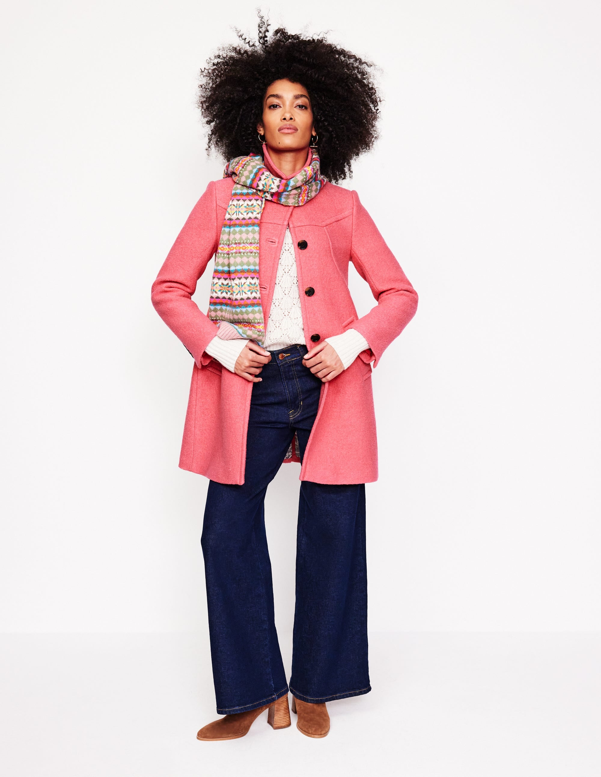 A woman wearing a textured Rhubarb-colored wool-blend coat with a neat collar and functional pockets, paired with a colorful scarf, dark blue jeans, and brown boots.